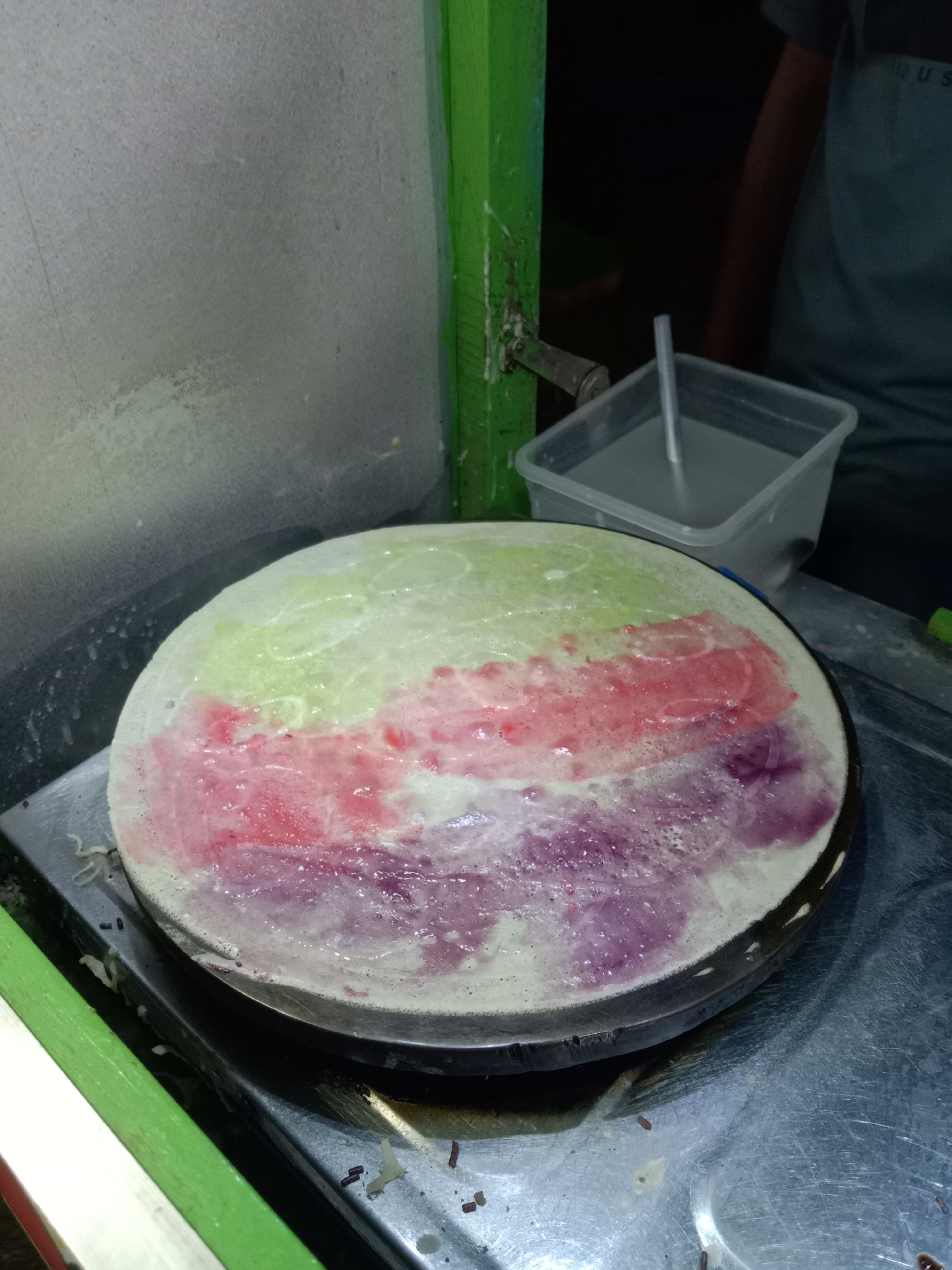 A close-up, high-angle shot shows a wide, circular crepe being cooked on a flat, circular griddle. The crepe batter is a pale, almost white color, and it is adorned with three distinct horizontal bands of colored toppings: a light green band at the top, followed by a pink band, and a purple band at the bottom. The toppings appear to be in a liquid or semi-liquid state, with some bubbling and spreading evident. The griddle is situated on a metal surface, possibly part of a food stall, and a portion of a green wooden frame is visible in the foreground, to the left of the griddle. In the background, a clear plastic container filled with a light-colored liquid and a metal utensil is visible, along with the arm and shoulder of a person wearing a dark t-shirt. The lighting suggests an outdoor or dimly lit environment, possibly at night. This scene is located in Jumput, Indonesia.