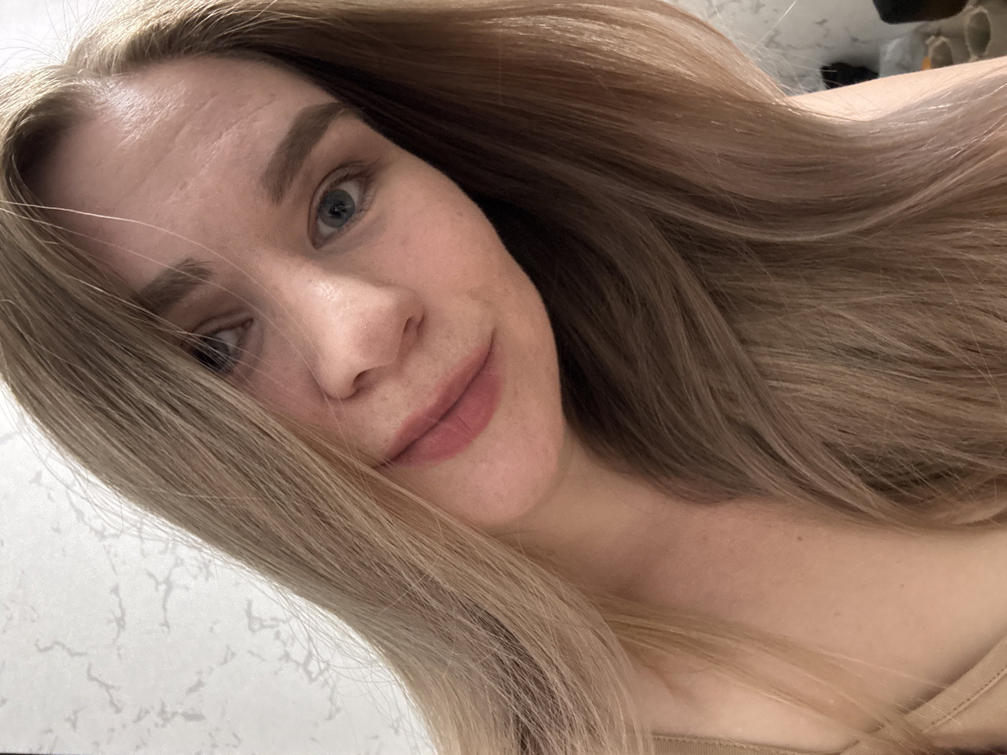 A close-up selfie shows a fair-skinned woman with long, wavy, light brown hair. Her face is angled towards the camera, with a slight smile. She has blue eyes and light eyebrows. The background is blurred and appears to be a white surface, possibly a wall or countertop. There's no indication of the city of Chaikovsky, Russia in the provided image.