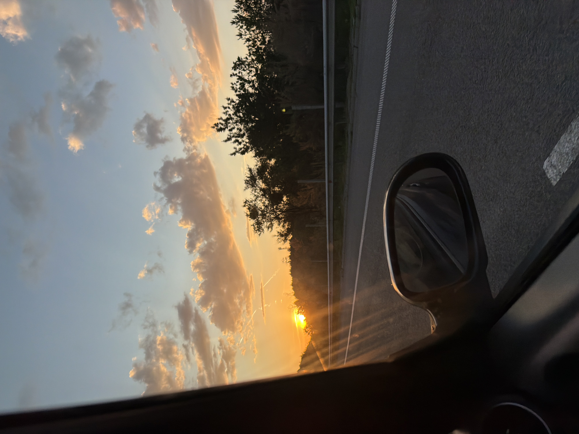 The image shows a sunset viewed from inside a car, likely driving on a road near Krushovitsa, Bulgaria. The view is from the driver's side, with the side mirror visible. The sky is filled with clouds illuminated by the setting sun. There are trees along the side of the road. The road surface has lane markings. The interior of the car is visible in the foreground.