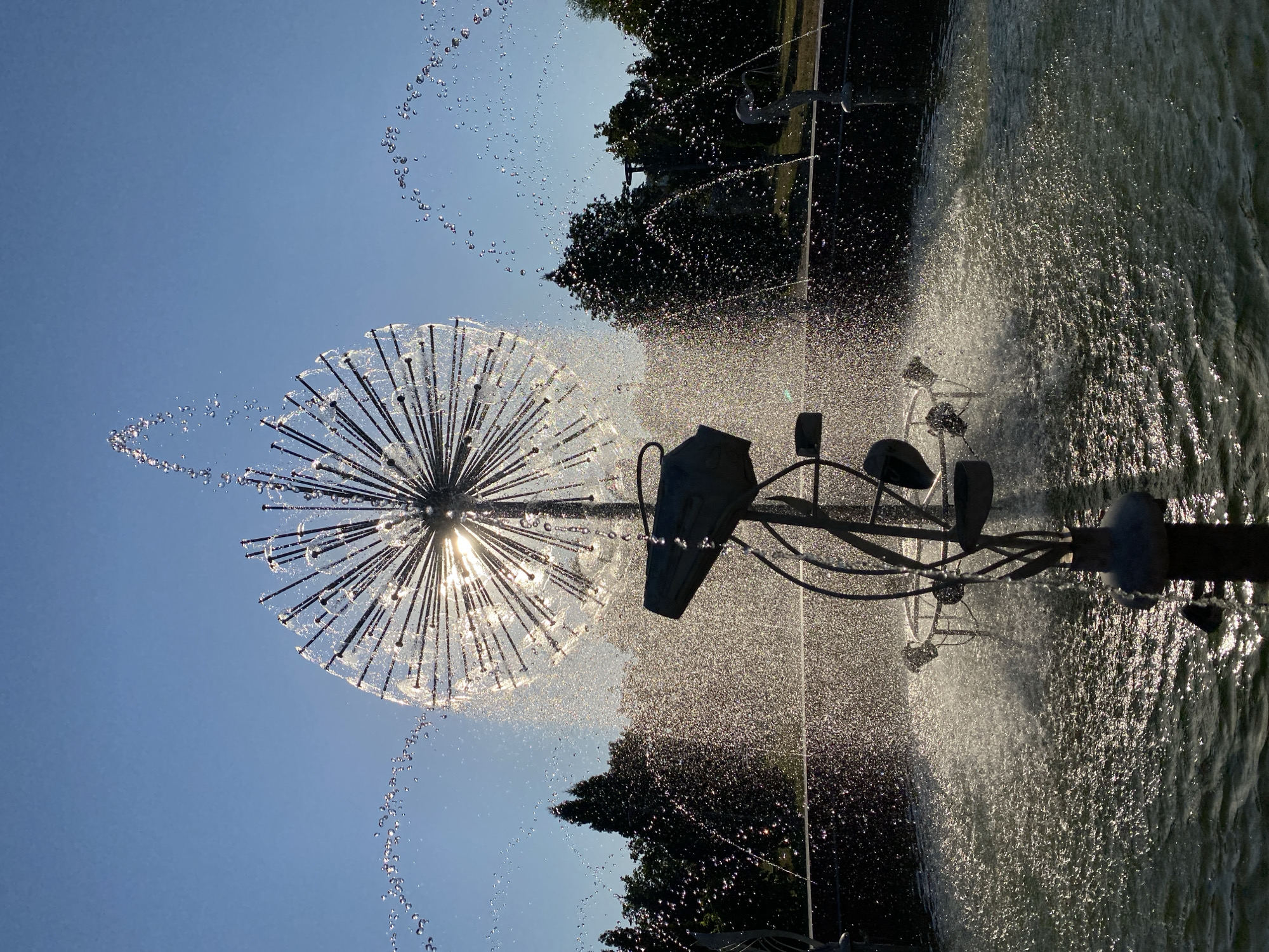 The image shows a dandelion-shaped fountain in a park. The fountain is made of metal rods and sprays water in all directions. The sun is shining brightly through the fountain, creating a halo effect. In the background, there are trees and a clear blue sky. The fountain appears to be located in a public park, likely in an urban environment. The specific location in Chișinău, Moldova is not identifiable from the image alone. The scene depicts a serene and refreshing outdoor setting on a sunny day.