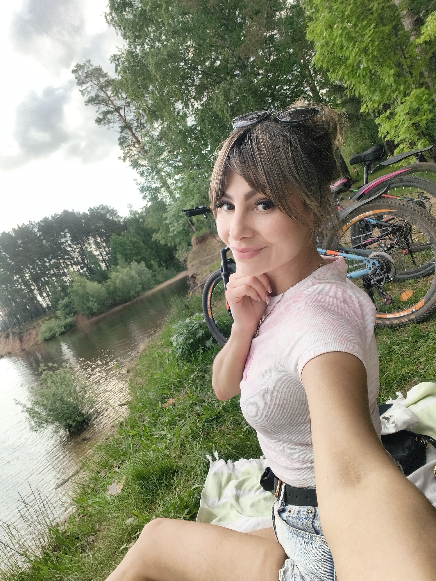 A woman is taking a selfie near a body of water. The woman has brown hair with bangs and is wearing sunglasses on her head, a pink tie-dye shirt, blue jean shorts, and a black belt. She is sitting on a blanket on the grass. The background shows a body of water with trees lining the shore. There are two bicycles parked behind her. The location is the city of Chaikovsky, Russia.