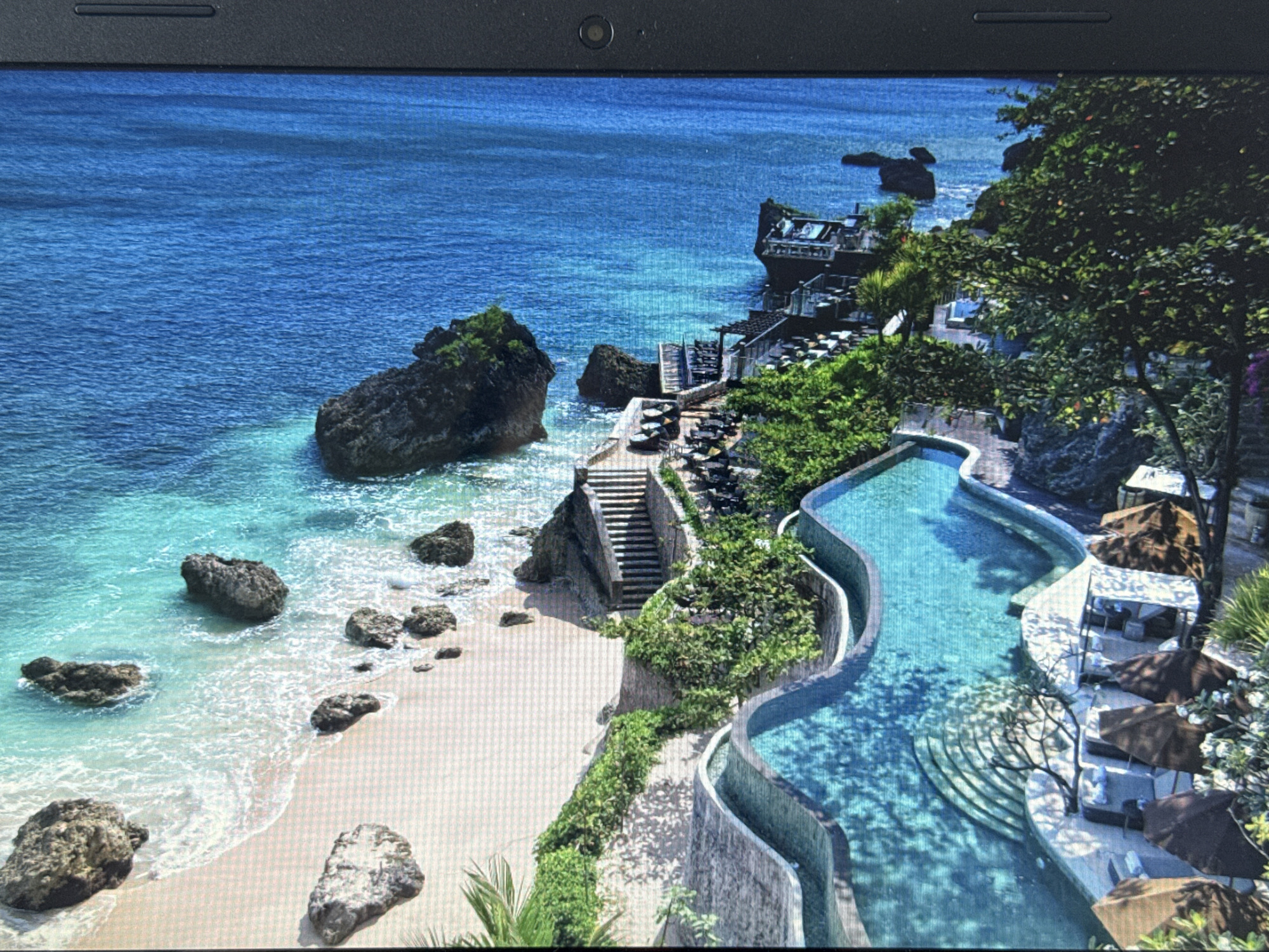 This image depicts a picturesque tropical resort situated on a cliff overlooking a vibrant blue ocean. The resort features a multi-level infinity pool with curved edges, surrounded by lush green vegetation and lounge chairs with umbrellas. A sandy beach with large rocks is visible at the base of the cliff, with gentle waves lapping at the shore. The resort itself appears to be built into the cliffside, with multiple terraces, outdoor dining areas, and a structure resembling a restaurant or bar perched on the edge. The architecture is modern and integrated with the natural landscape. The scene is bathed in bright sunlight, highlighting the clear blue of the water and the vibrant green of the foliage. There are no people visible in the image. The image is presented on a laptop screen, with the edge of the laptop and its webcam visible at the top of the frame. The city of Pushkino, Russia is not depicted in this image.