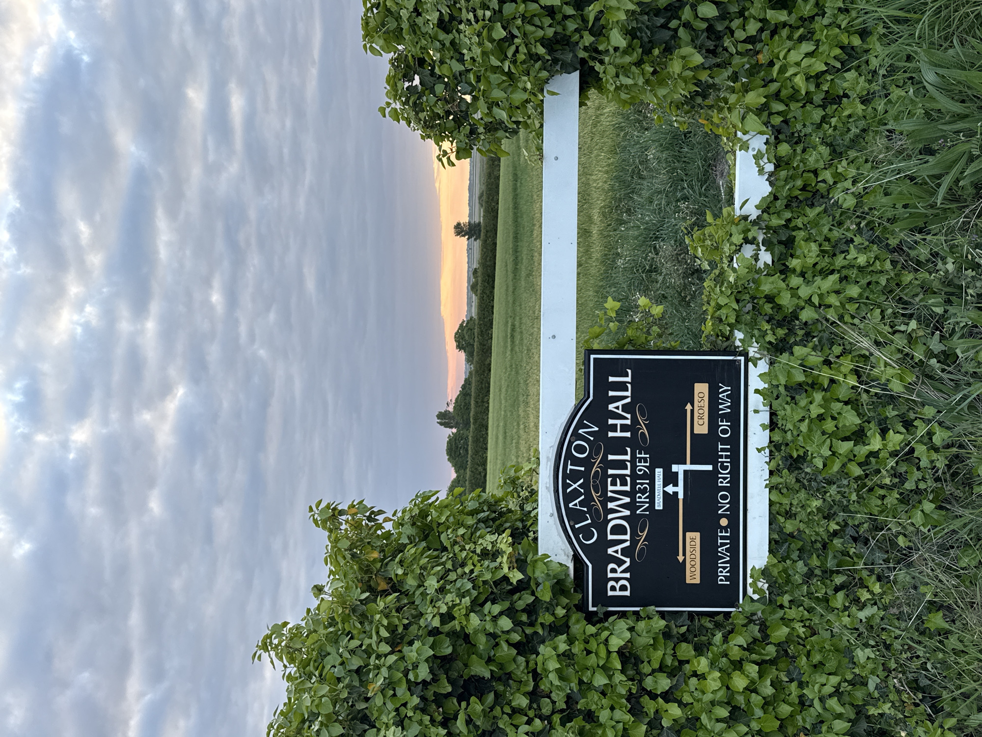 The image depicts a black and white sign located outside Claxton Bradwell Hall, NR31 9EF, which is in the city of Great Yarmouth, United Kingdom. The sign is partly obscured by green foliage, likely ivy. The sign reads "Claxton Bradwell Hall" and "Private No Right of Way" with directional arrows indicating "Woodside" and "Croeso". The sign is attached to a white post. Beyond the sign, there is a view of a green field under a sky with cloud formations. The light indicates sunset or sunrise.