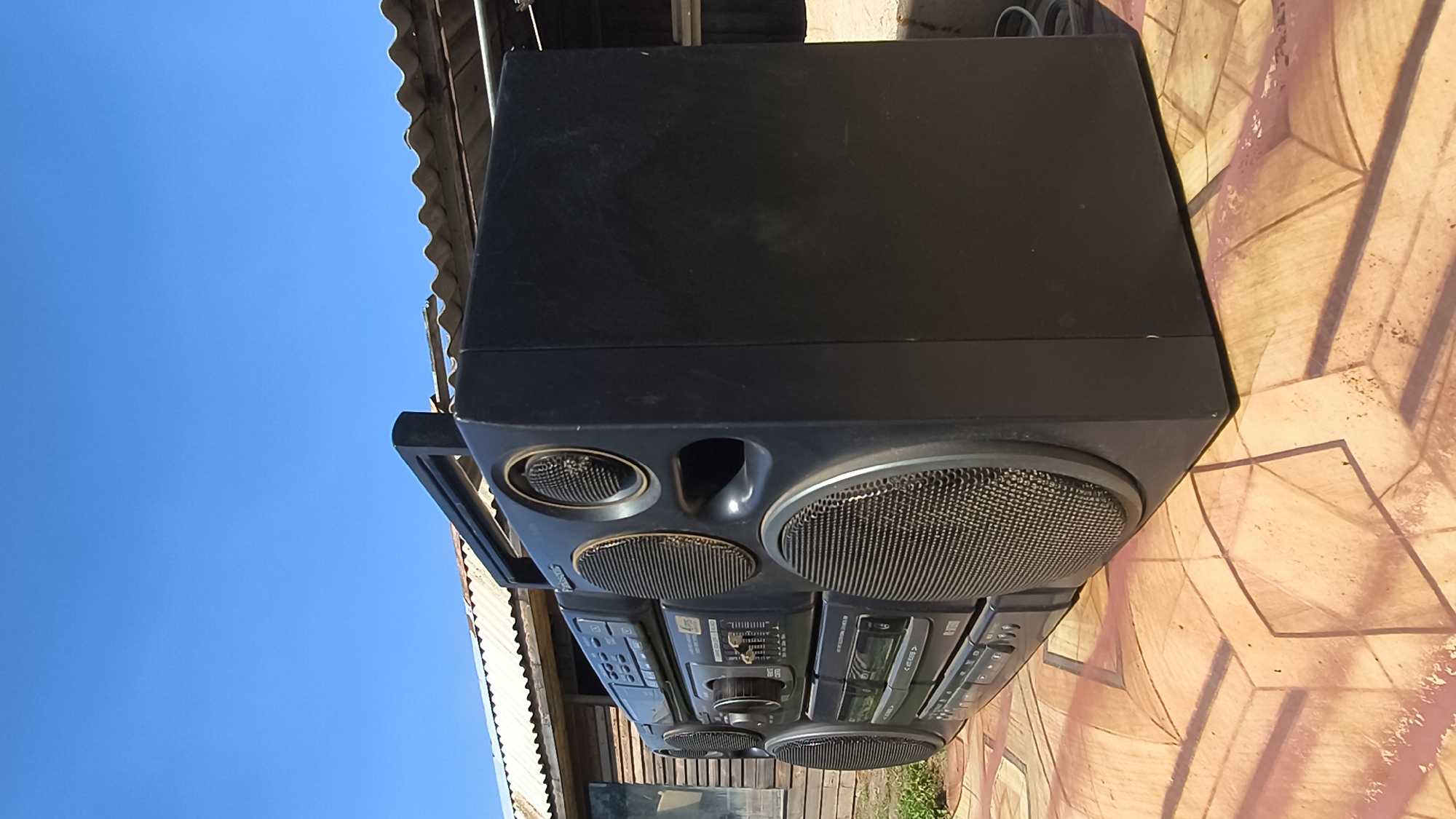 An outdoor, eye-level, medium shot captures a black Panasonic boombox placed on a patterned surface under a clear blue sky. The boombox is angled from the left side, with its right side facing the viewer. It features multiple speakers, buttons, and a cassette player. Behind the boombox, a weathered wooden building with a corrugated metal roof is visible. The patterned surface appears to be a tiled or painted ground with geometric designs. The scene is devoid of people. The setting is likely a yard or outdoor area in Mikhaylovka, Russia, given the overall aesthetic and architectural elements.