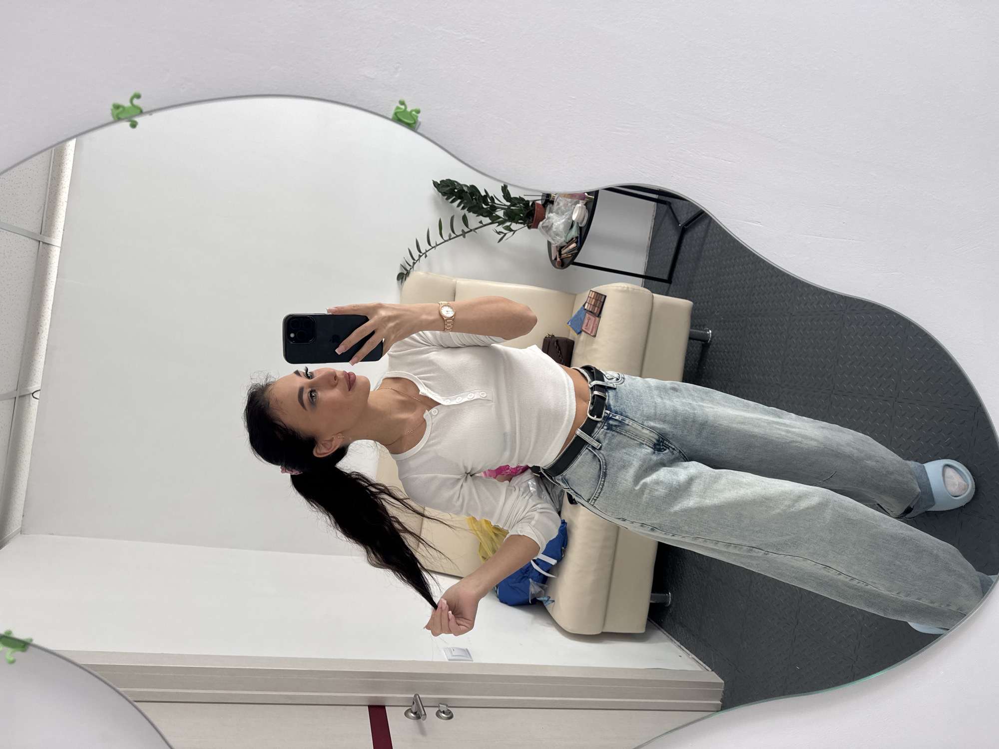 A woman is reflected in a large, oddly shaped mirror. The mirror's frame is decorated with small green frog figurines. The woman is holding a cell phone, presumably taking a selfie. She has long, dark hair pulled back into a ponytail and is wearing a white, long-sleeved crop top, light-wash jeans, a black belt, and light blue slippers. Behind her is a beige loveseat with a plant on a black table to the side. The floor has a diamond-plated pattern. The background suggests an indoor setting, possibly an apartment or dressing room. The city of Фоки, Russia, is not discernible from the image.