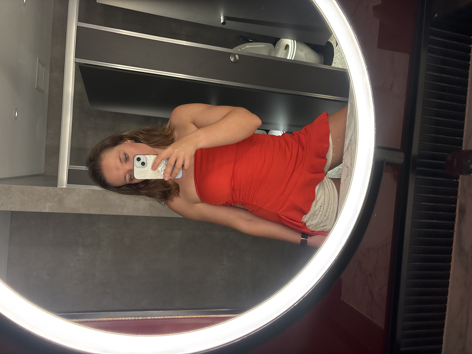 A woman is taking a mirror selfie in a restroom. She is wearing a red strapless top and white shorts. The restroom has grey tiled walls and a white illuminated mirror. A toilet is visible in the reflection of the mirror. The image does not contain information to determine the location in the city of Đà Nẵng, Vietnam.