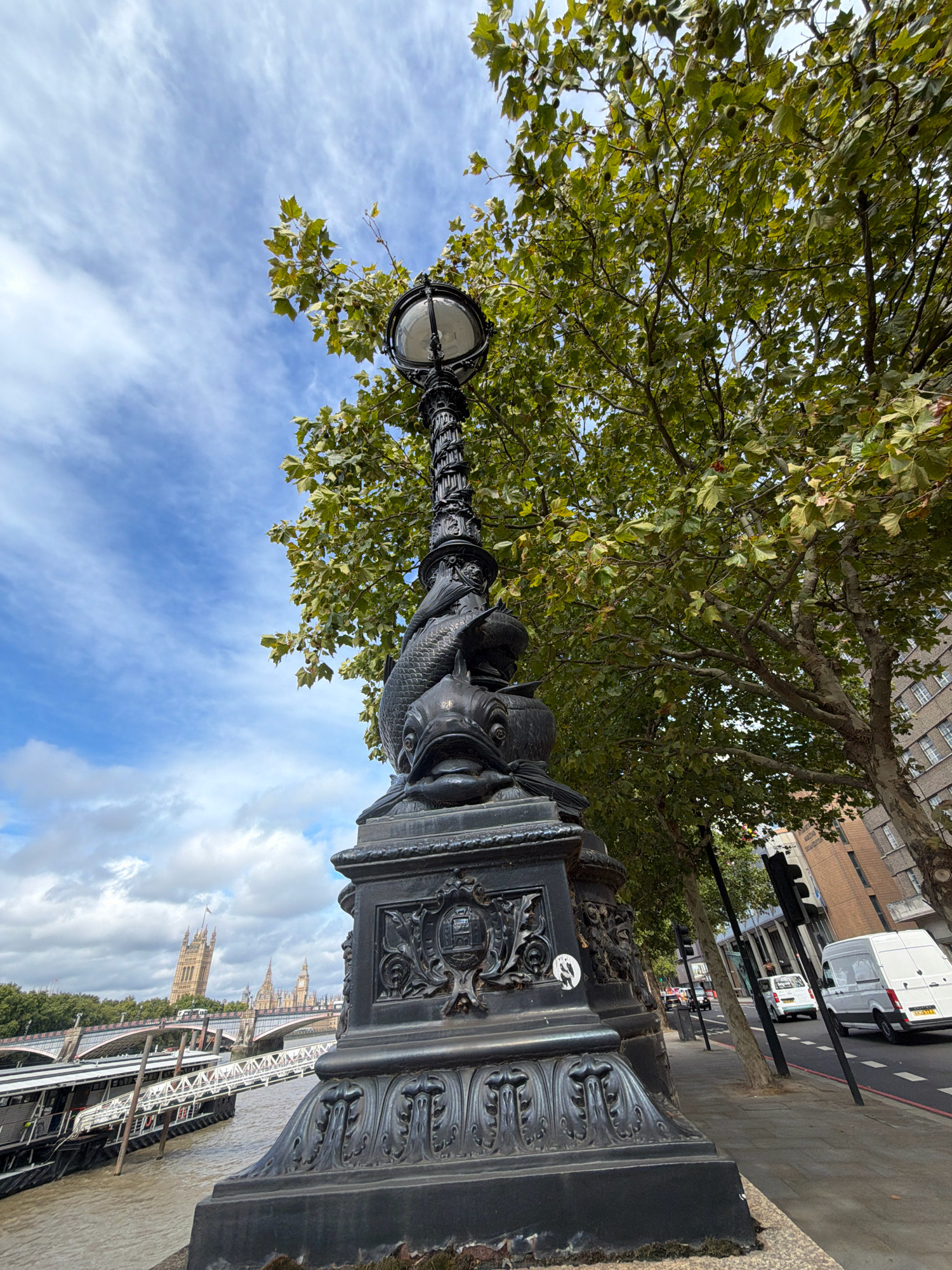 The image captures a low-angle view of an ornate, black lamppost with a fish-like design at its base. The lamppost is positioned on a sidewalk next to a tree with green foliage. In the background, across a body of water, the iconic Houses of Parliament and Big Ben are visible, indicating the location as London, United Kingdom. The Thames River is in the foreground, with a boat docked beside it. The sky is partly cloudy. A street with vehicles and buildings lines the sidewalk.