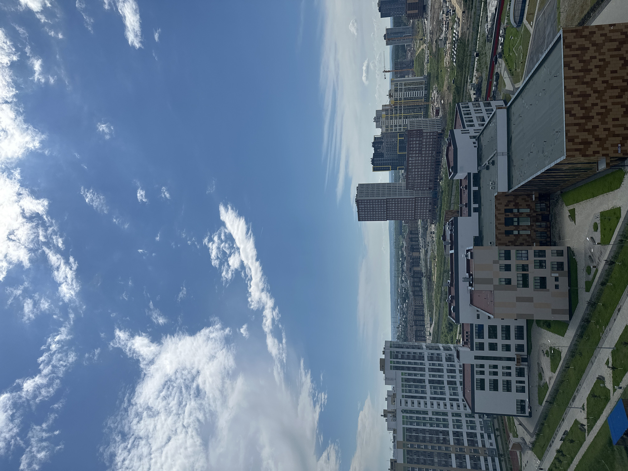 This image captures a modern urban landscape in Yekaterinburg, Russia, featuring a collection of contemporary residential and commercial buildings under a bright blue sky with scattered clouds. The foreground is dominated by the architecture of several multi-story buildings, characterized by their geometric designs and varied facades, some with large windows and balconies, others with distinctive patterned claddings. Green spaces with manicured lawns, pathways, and benches are visible around the buildings, indicating a planned urban development with recreational areas. In the mid-ground, a cluster of taller buildings, including high-rises, forms a city skyline. Construction cranes are visible among some of these structures, suggesting ongoing development. The background reveals a more distant urban expanse with rows of buildings and hints of natural terrain beyond. The scene is devoid of prominent human activity, focusing instead on the architectural structures and the expansive sky.