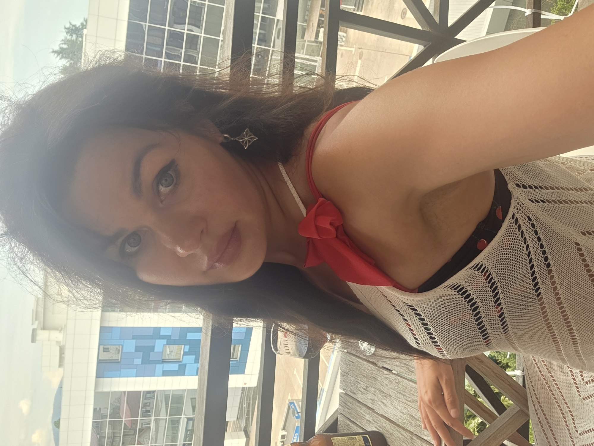 A woman is taking a selfie on a balcony overlooking buildings in Sochi, Russia. She has dark hair and is wearing a white open-knit cover-up over a red and black polka dot bikini top. A red bow detail is visible on the left strap of her bikini. She is also wearing a small, star-shaped earring. In the foreground, a wooden table holds a bottle of beer and a wine glass filled with red liquid. The background shows modern buildings with reflective windows, typical of Sochi. The sky is overcast.