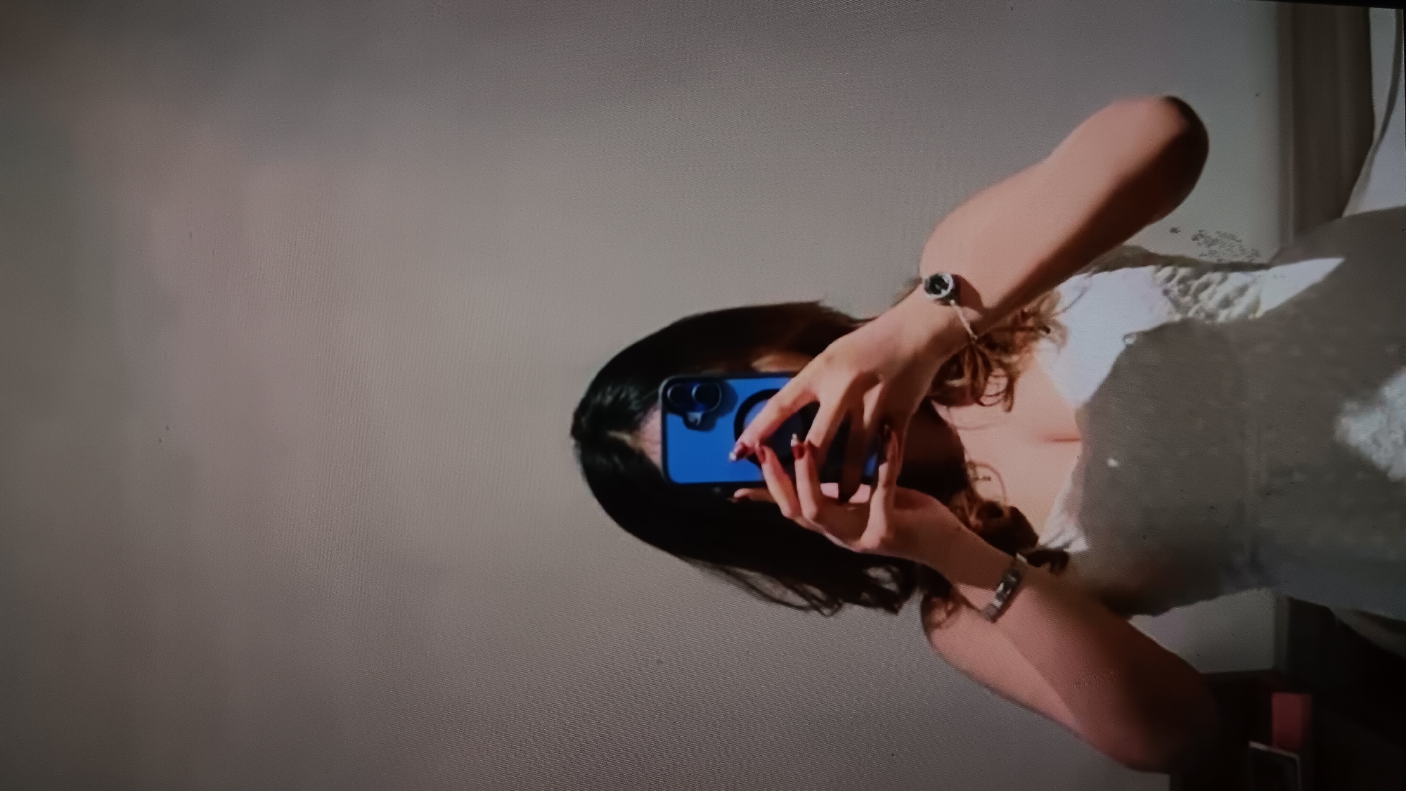 A young woman is taking a mirror selfie in a room, possibly a bedroom or dressing room. She is wearing a white, strapless dress that appears to be made of lace or a similar textured fabric. Her hair is dark and styled loosely, falling around her shoulders and face, partially obscuring her features as she holds a bright blue smartphone in front of her face to capture the photo. Her fingernails are painted red. On her left wrist, she wears a wristwatch with a dark face and a silver band. The background is a plain, light-colored wall, possibly textured. To the left of the woman, a portion of a dark rectangular object, possibly a piece of furniture or a box, is visible on a lower surface. The lighting in the room is dim, creating shadows that add to the intimate and personal nature of the image. The overall impression is a casual, self-portrait taken in a private space, suggesting a moment of personal reflection or preparation. The image does not contain any elements that would indicate a specific location within North Jakarta, Indonesia.