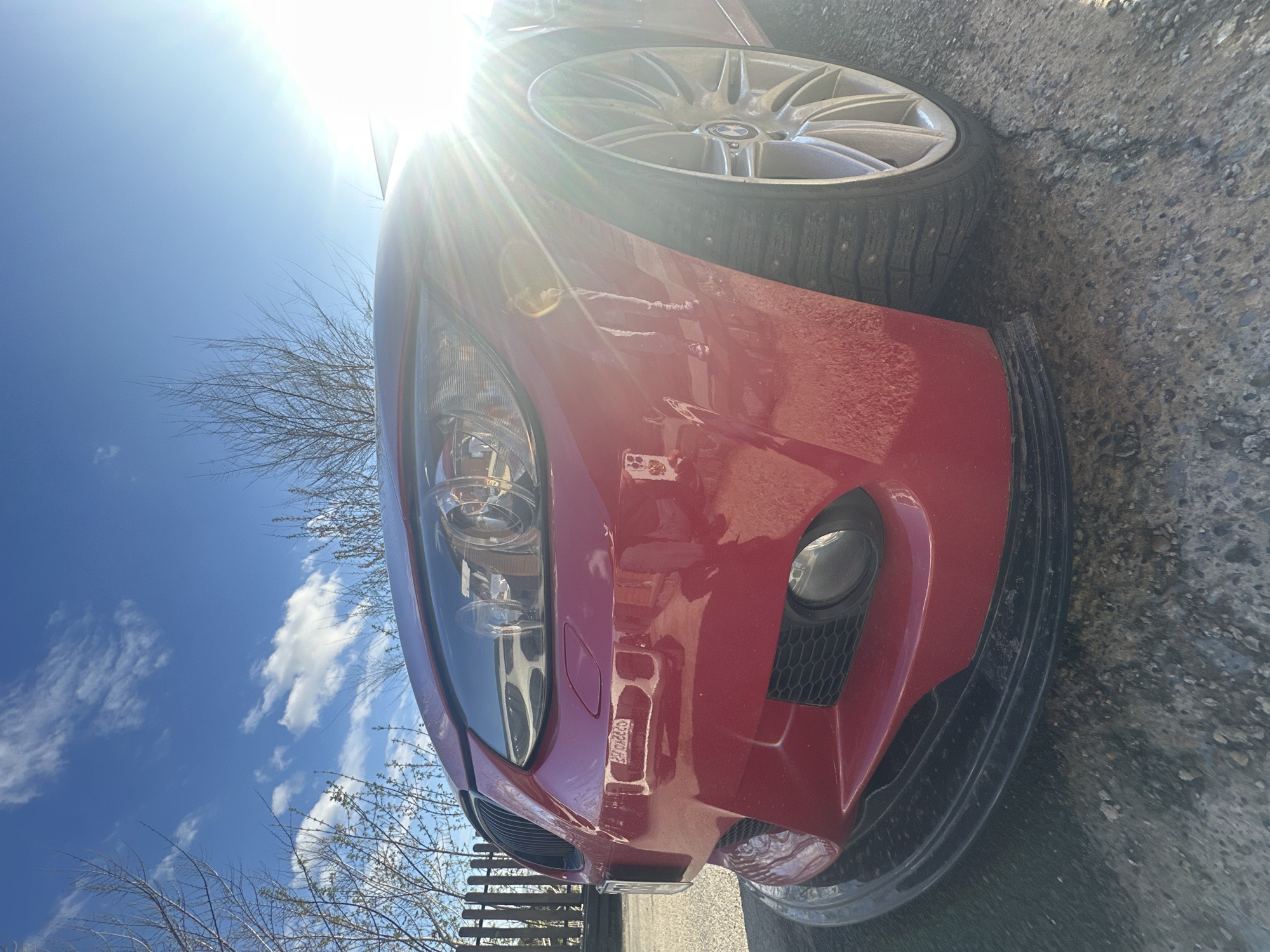 The image shows the front end of a red car parked on an asphalt surface. The car is a BMW, as evidenced by the emblem on the silver wheel. The tire is studded. The sky is visible in the background, showing a clear blue with some white clouds. There are bare tree branches also in the background, near what may be a fence. The image is taken in bright sunlight, causing lens flare. The location is Unknown, Unknown.