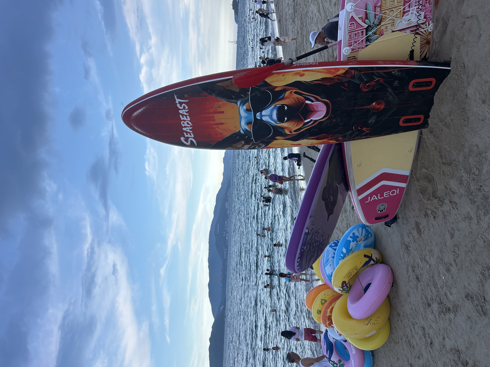This image shows a beach scene in Nha Trang Ward, Vietnam. In the foreground, several inflatable rings are piled on the sand, with a stand-up paddleboard featuring a graphic of a dog wearing sunglasses in the center. Behind these objects, the ocean is visible with numerous people swimming and wading. In the distance, hills with buildings are silhouetted against a cloudy sky. The overall scene depicts a popular beach destination with recreational activities.