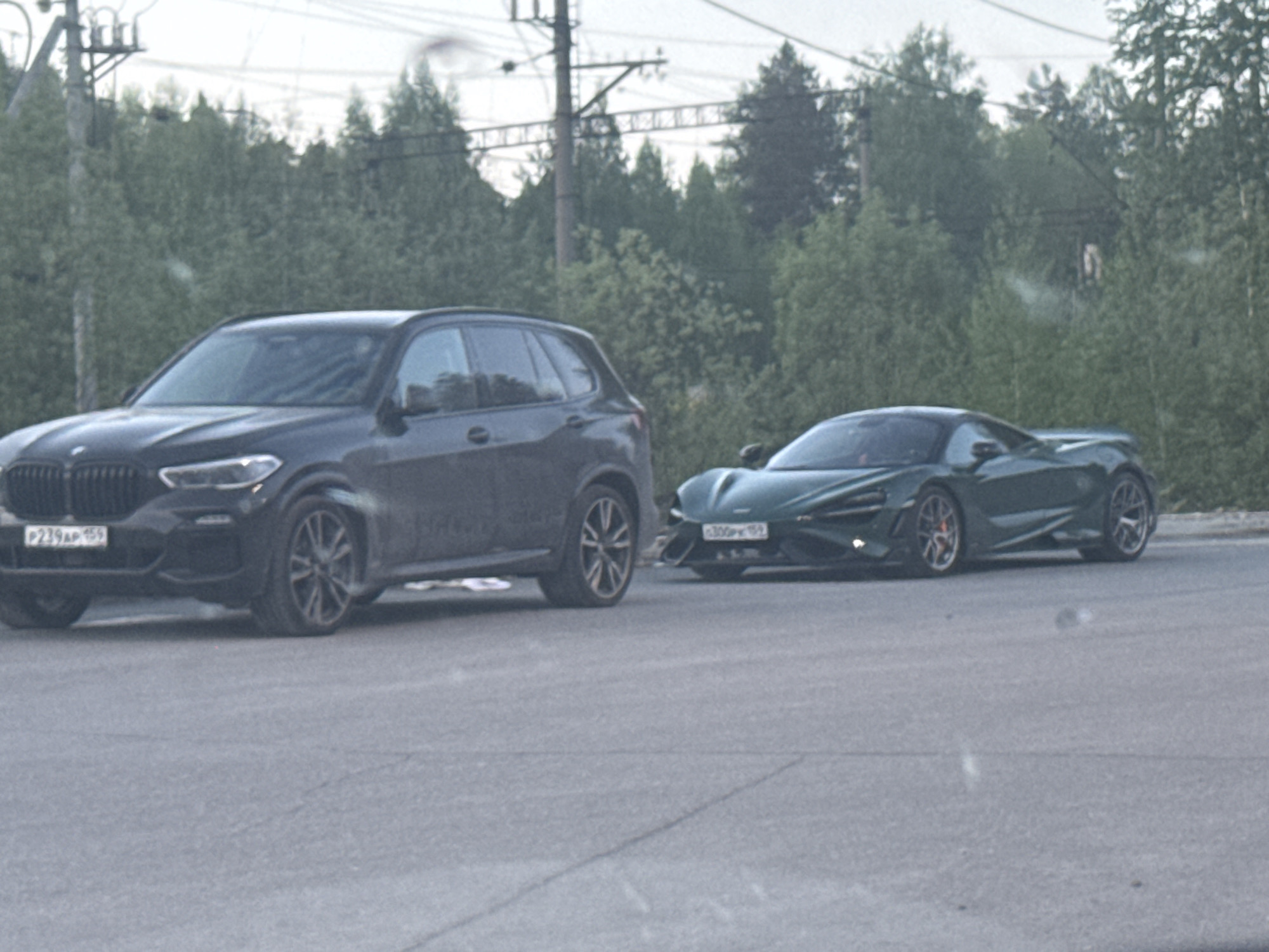 The image shows a black BMW X5 and a green McLaren 720S parked on a paved road. The BMW has a license plate "P239AP 159". The McLaren has a license plate "C300PX 159". In the background, there are trees and utility poles. The image was likely taken in Perm, Russia based on the license plates that indicate the 159 region.