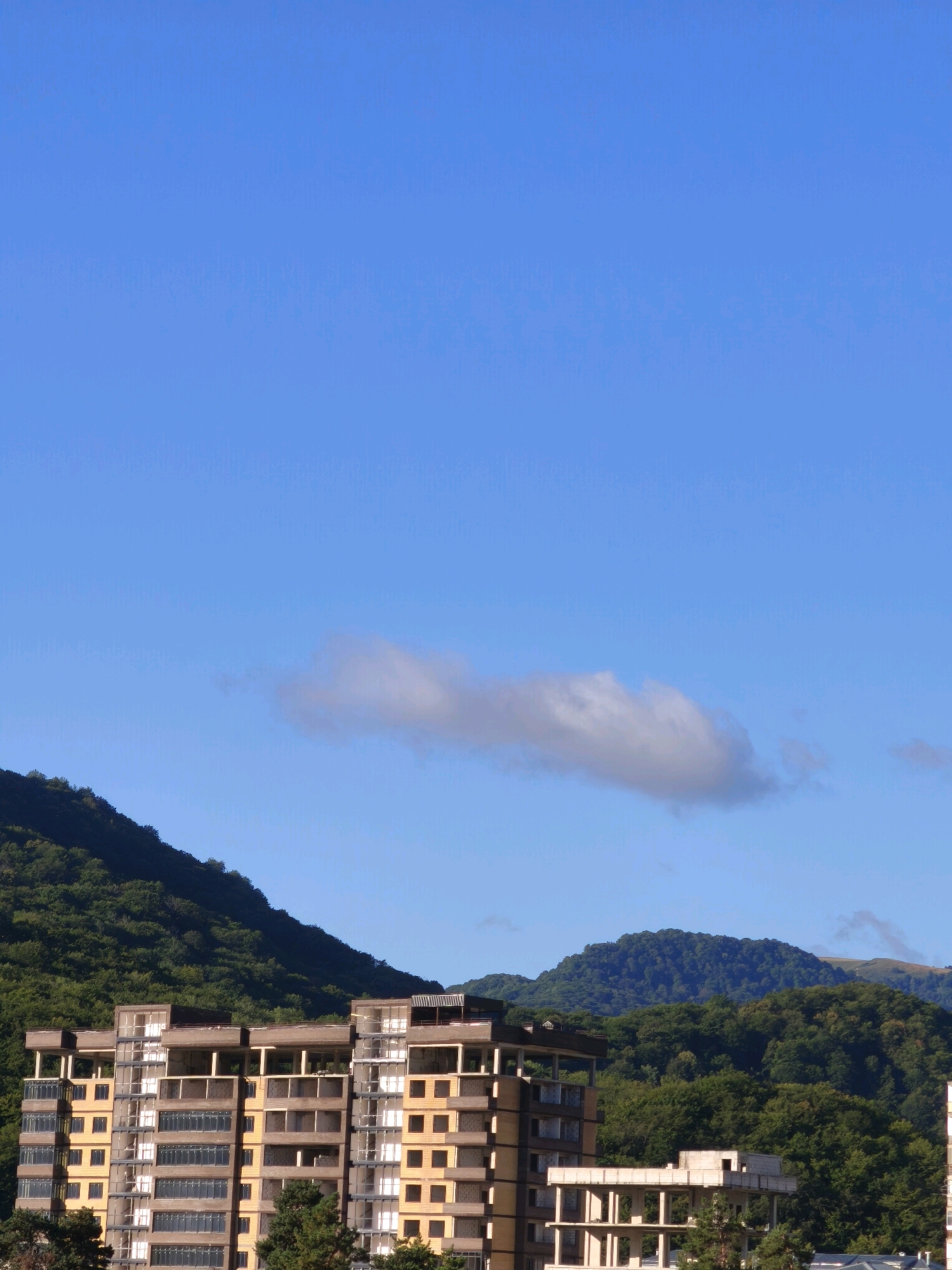 An unfinished concrete building stands in the foreground against a backdrop of lush green mountains under a clear blue sky with a single wispy cloud. The building, which appears to be a residential complex, has multiple floors with exposed concrete structures, balconies, and many empty window frames. The surrounding natural landscape suggests a location with significant elevation and dense vegetation. This scene is likely located in the city of Karachayevsk, Russia, known for its mountainous terrain and opportunities for outdoor recreation.