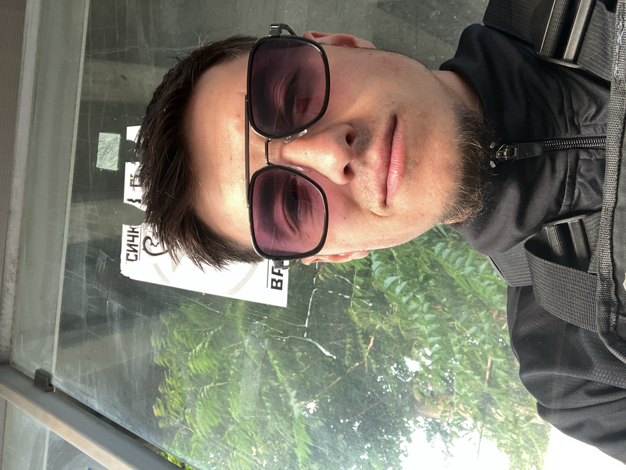 A man is photographed inside a bus stop in Plovdiv, Bulgaria. He is wearing sunglasses and a black jacket with a backpack. The bus stop has a glass window with some stickers on it. Trees and foliage are visible through the glass.