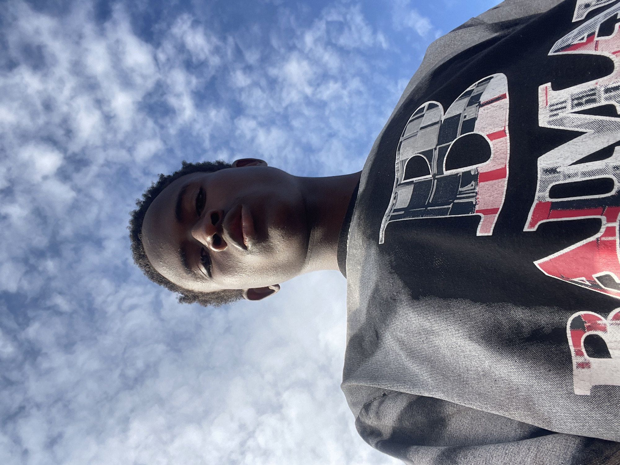 A low-angle, wide shot shows a young Black male, viewed from the chest up, against a bright blue sky with scattered white clouds. He is looking directly into the camera with a neutral expression, his lips slightly pursed. His hair is short and dark. He is wearing a dark crewneck sweatshirt with a large graphic design on the front. The design features a large white letter "B" filled with a plaid pattern in black, grey, and red. Below the "B", the letters "AMA" are visible in white, also filled with a similar plaid pattern. The sweatshirt appears to be made of a textured fabric. The background is entirely the sky, suggesting the photo was taken outdoors during the daytime. The angle of the shot and the bright sky suggest it might be midday. The image was captured in Jos, Nigeria.