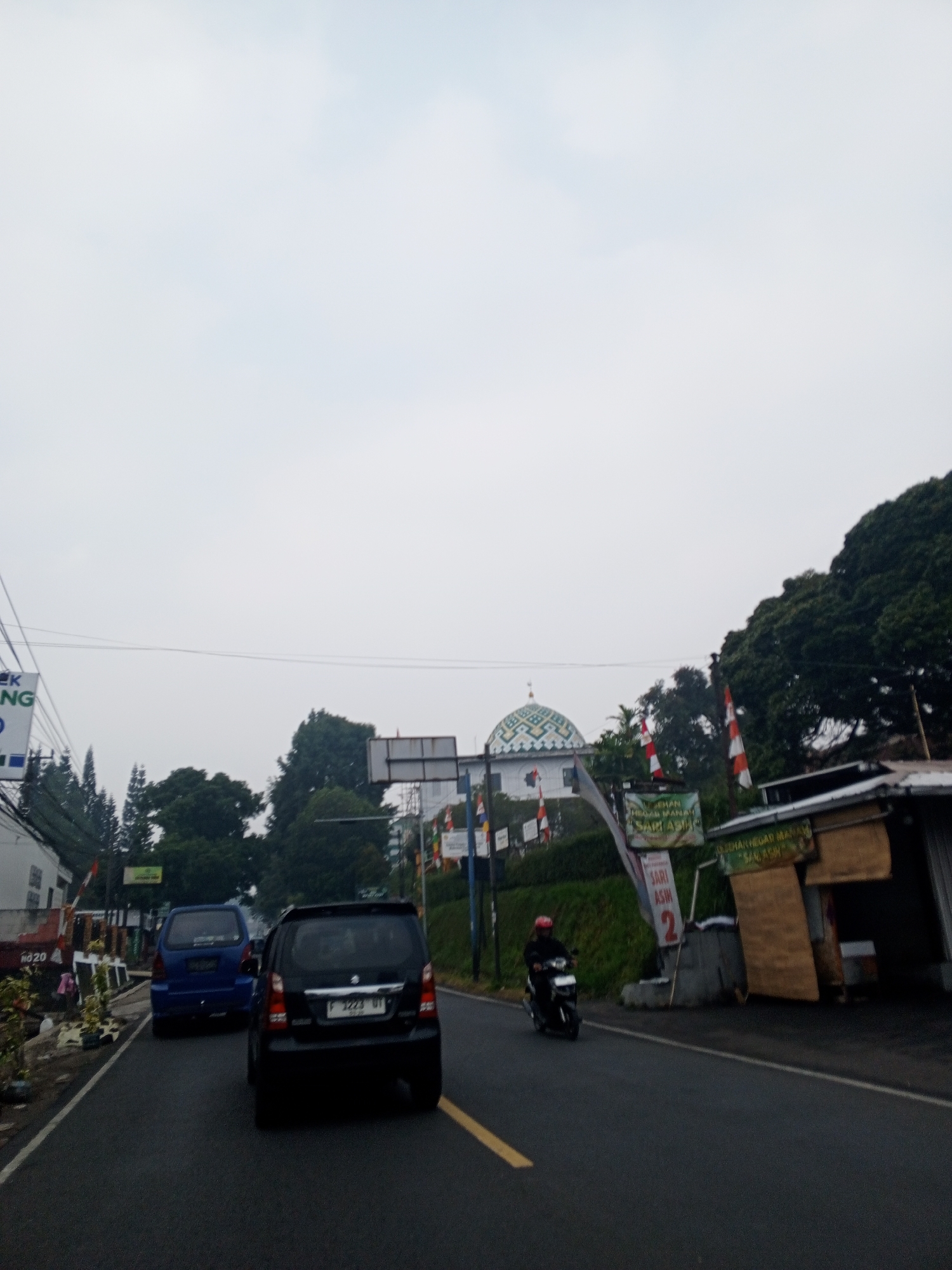 The image shows a street scene. In the foreground, there is a dark-colored car on the right side of the road and a blue car on the left side. A motorcycle with a rider in a red helmet is also visible on the road. In the background, there is a mosque with a green dome and white accents, flags, and trees. There are also some buildings and signs along the roadside. The sky is overcast. The image does not show anything that would indicate the location is Dambulla, Sri Lanka.
