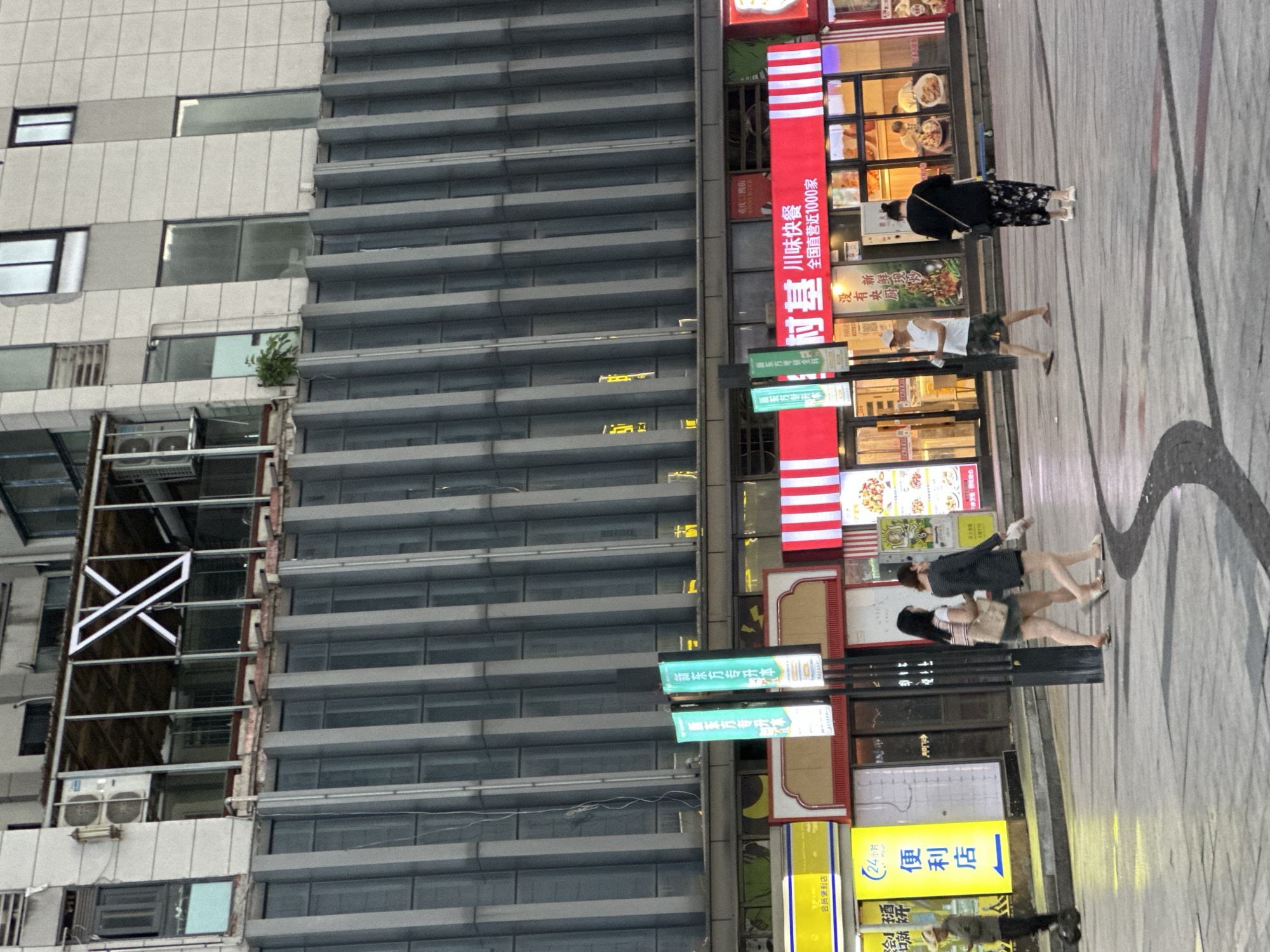 The image depicts a street scene in Chongqing High-tech Zone, China. The setting is urban, with a tall, modern building occupying the left side of the frame. The building's façade is a mix of grey panels and windows, with some sections appearing to be under construction or renovation, indicated by scaffolding and exposed building elements. Adjacent to the building are a series of commercial establishments or shops, distinguished by bright signage and displays. A prominent red sign reads "村基味快餐 全国直营近1000家" (Cunji Flavor Fast Food, nearly 1000 direct-operated stores nationwide), suggesting a chain restaurant. Additional signs indicate other businesses, including a convenience store, "便利店" (Convenience Store), and educational services, "新东方专升本" (New Oriental Bachelor's Degree Upgrade). Several people are present on the sidewalk, mostly pedestrians walking and seemingly engaged in typical urban activities. They are dressed casually in a mix of clothing styles. The ground is paved, with dark lines or patterns indicating possible walkways or traffic flow. The overall impression is that of a bustling commercial area within a modern Chinese city.
