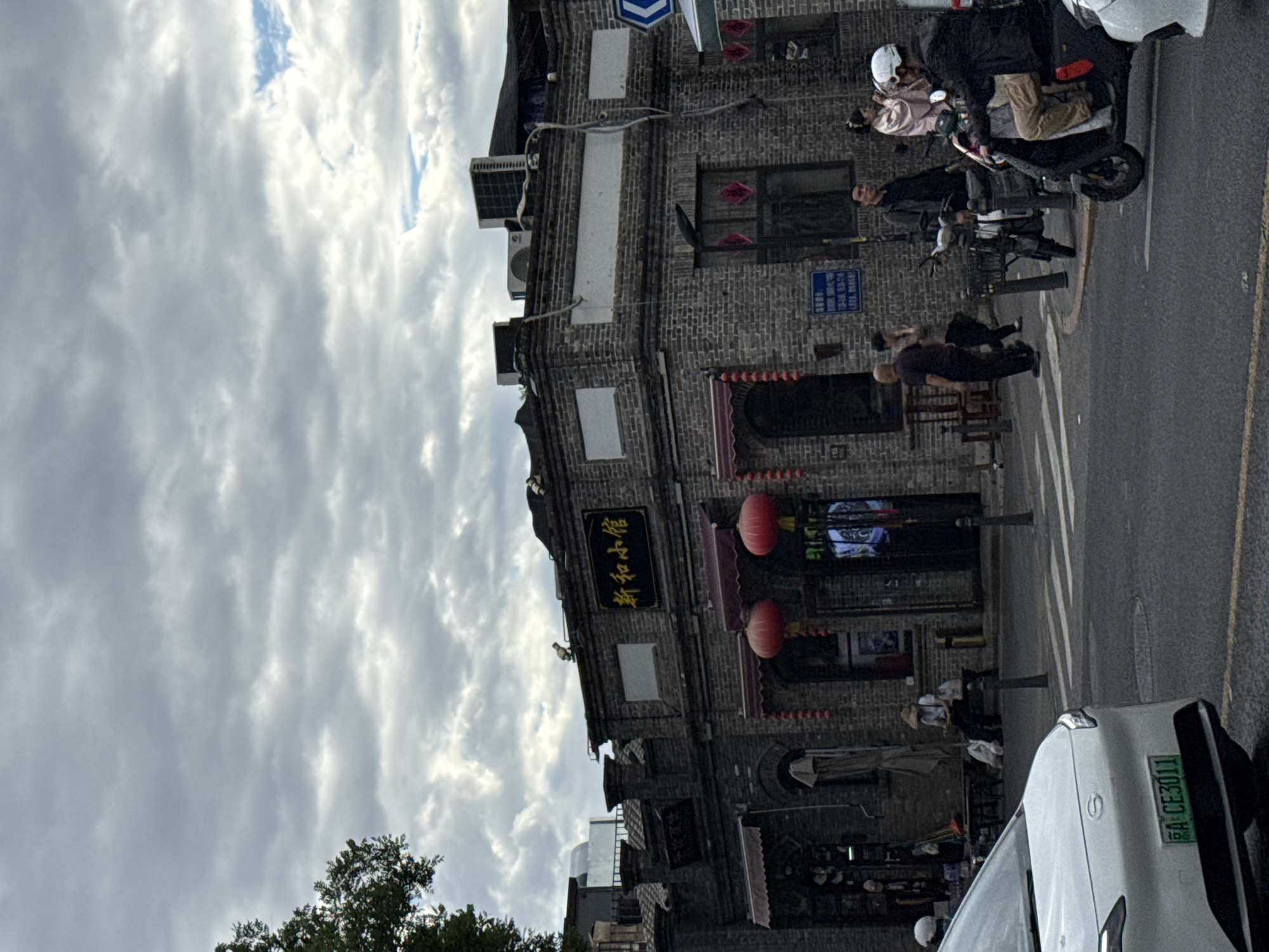 The image captures a street scene in Dongcheng District, China, under a cloudy sky. A two-story brick building, appearing to be a shop or restaurant with traditional architectural elements, is the central focus. Red lanterns hang from the building's facade, and a golden sign with Chinese characters reads "新和小馆" (Xīn Hé Xiǎo Guǎn). Several people are visible on the street. In the foreground, a white car with a license plate "EA CE3011" is parked. To the right, a person wearing a white helmet rides a scooter. Other individuals are walking or standing near the building. The street is paved, with crosswalk markings visible. The overall atmosphere suggests a commercial or residential area with a blend of old and new elements.