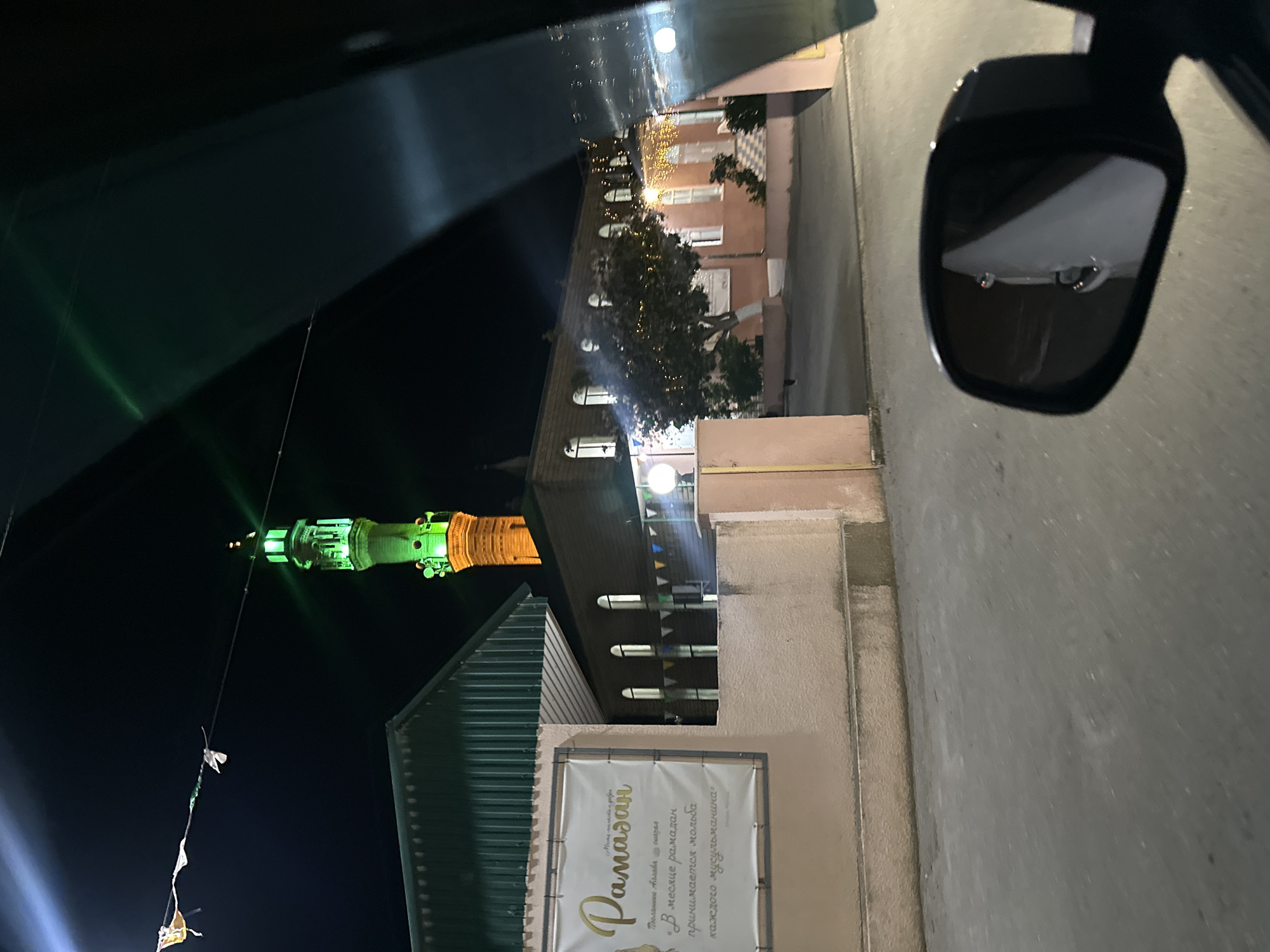 This is a vertical, nighttime photograph taken from inside a vehicle. The scene depicts a street with buildings and a brightly lit minaret of a mosque, illuminated in green and orange. Trees decorated with festive lights are visible between the buildings. In the foreground, a side-view mirror reflects a portion of the vehicle's interior and the street.
A banner with text is displayed on the side of a building. The visible text, written in Cyrillic and possibly Arabic script, appears to be related to Ramadan. The text reads "Ramadan" in large golden script, with smaller text below.
The overall impression is one of a festive evening celebration, likely during the month of Ramadan, in a town with a prominent religious structure. The lighting suggests it is after sunset, and the presence of decorative lights implies a celebratory atmosphere. The reflection in the mirror adds a sense of perspective, indicating the image was captured while the viewer was in motion.