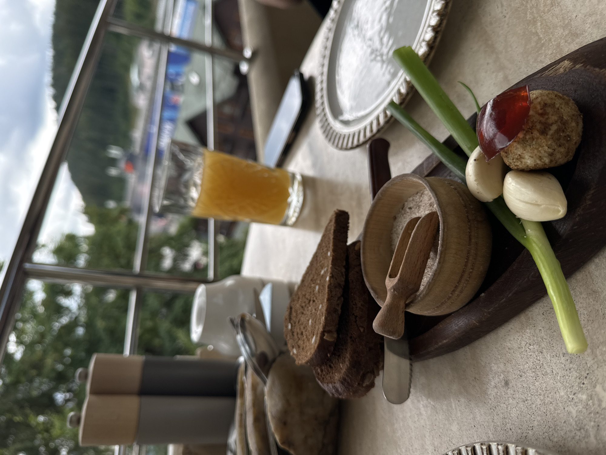 A table setting on a balcony in Polianytsia, Ukraine, features several food items and beverages. In the foreground, a wooden board holds a small wooden bowl filled with granular seasoning, a wooden scoop, two cloves of garlic, a small, round food item topped with a red jelly, and two stalks of green onion. To the left, a butter knife rests on the board, next to two slices of dark bread on top of a larger loaf. A glass of orange juice sits in the background, next to a mobile phone and a white ceramic container. Two salt and pepper grinders are visible to the far left. The balcony railing and lush green trees create a backdrop, suggesting an outdoor dining environment.