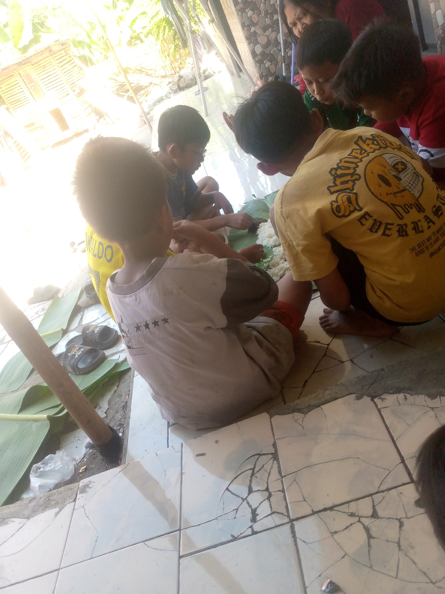 In Giriasih, Indonesia, several children are gathered on a tiled floor, sitting on banana leaves, and eating. The scene appears to be outdoors, with natural light illuminating the area from the left. The children are focused on the food, which is piled on the leaves in front of them. One child, wearing a yellow shirt with a skull graphic, is particularly prominent, facing away from the camera. Another child in a light-colored shirt is also visible, with their back to the viewer. Other individuals, including an adult, are partially visible in the background. The environment suggests a simple, communal setting, possibly a home or outdoor eating area. The tiled floor is cracked in several places. A wooden tool and a plastic bag are also present on the ground.