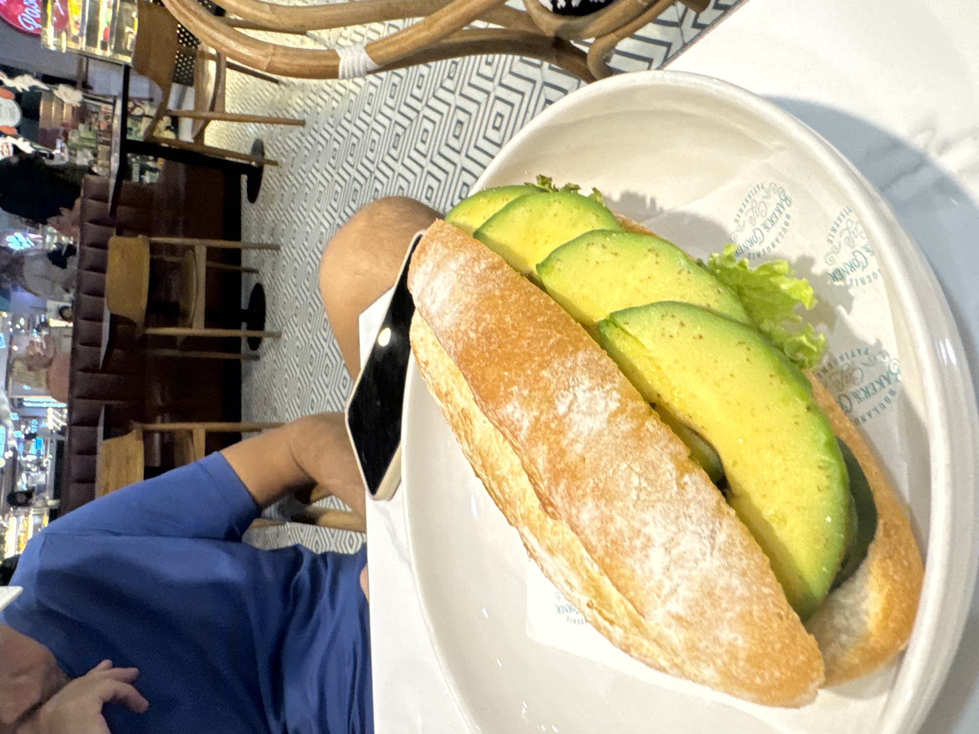 A close-up, eye-level shot shows a person's arm and torso in the foreground, wearing a blue t-shirt. They are seated at a white table in a cafe in Kuta Selatan, Indonesia. In the center of the frame, a white bowl holds a baguette sandwich filled with sliced avocado and lettuce. The sandwich is on a white paper liner with a faint green logo. The background shows cafe seating with wooden chairs and booths, as well as other patrons. The floor has a geometric tile pattern.