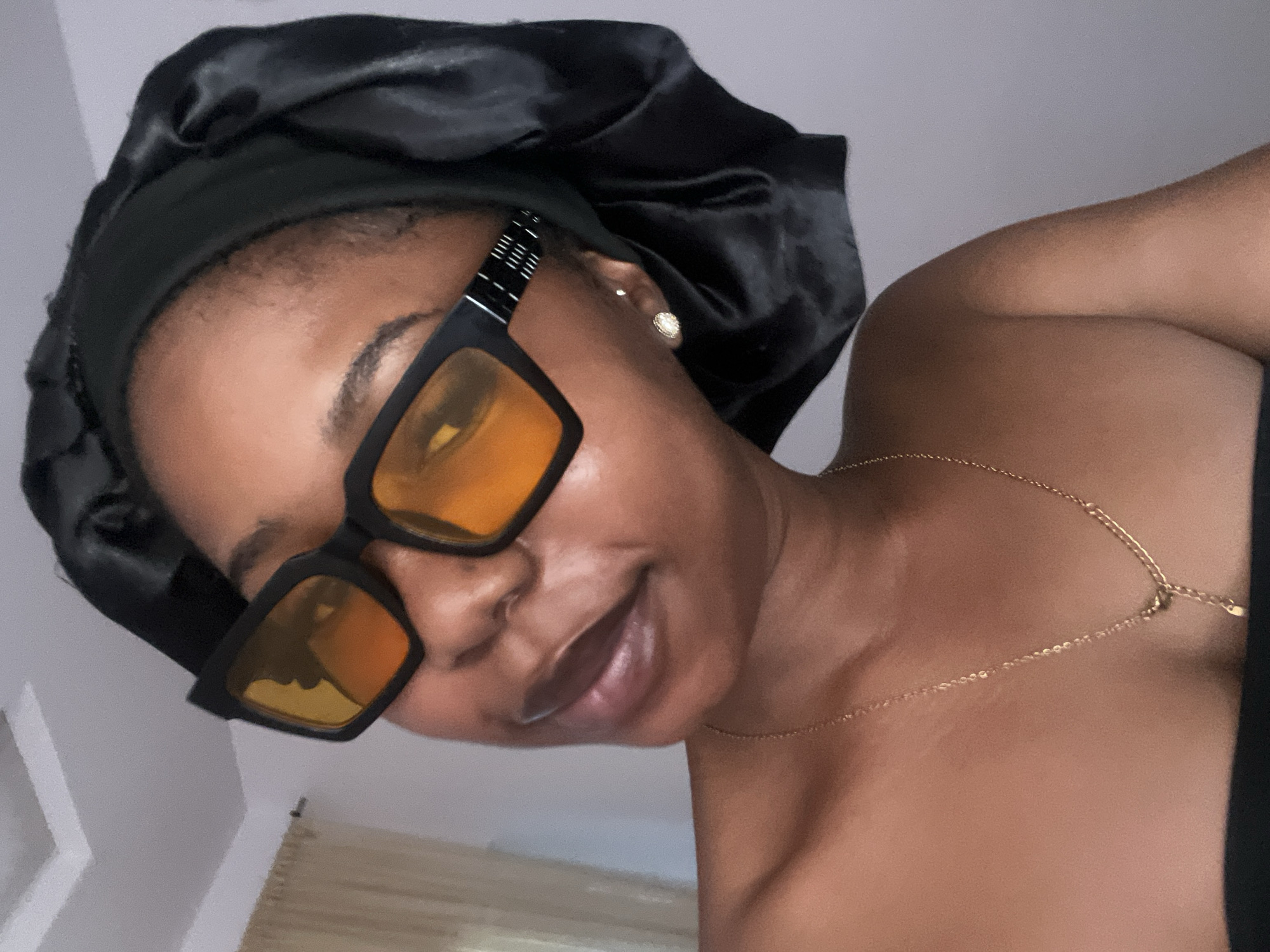 A young Black woman is captured in a close-up, head-and-shoulders shot. She is wearing a black satin bonnet, a gold necklace, and black-rimmed sunglasses with orange lenses. Her left shoulder and upper chest are visible, and she appears to be indoors, with a light-colored ceiling and curtain in the background. The lighting suggests natural light from a window. The image focuses on her facial features, with her head tilted slightly. The overall impression is a casual, self-portrait. The specific location of Rafin Par, Nigeria, is not discernible from the visual information in the image.