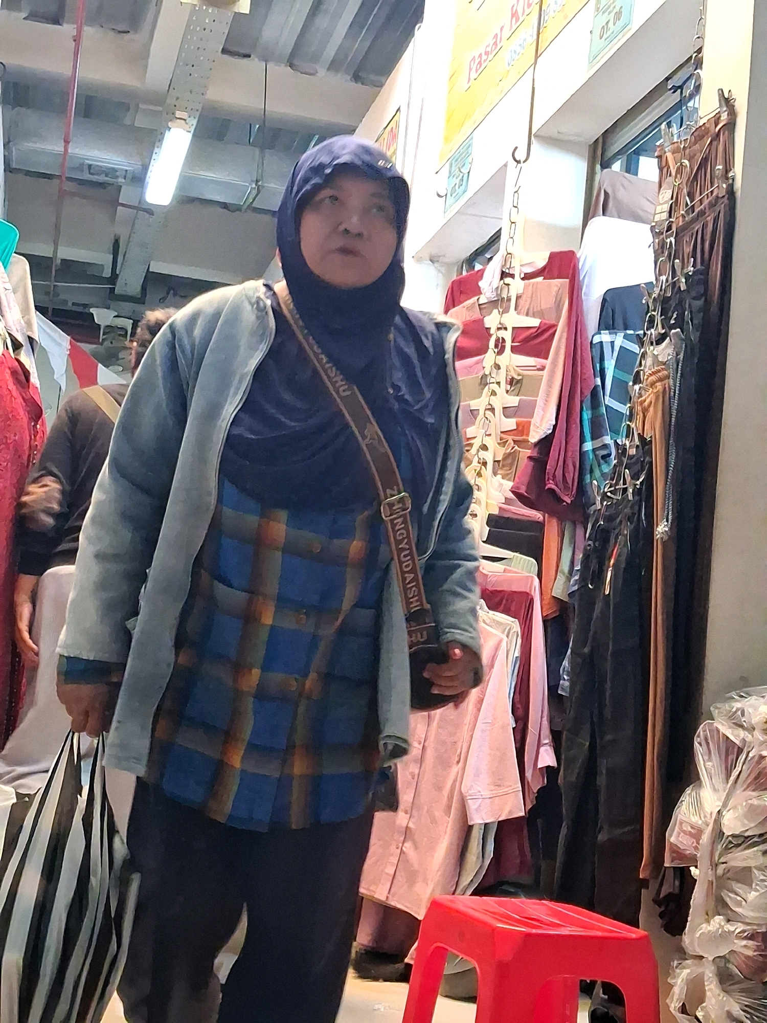 A woman wearing a dark blue hijab and a light blue jacket over a blue and orange plaid shirt walks through a crowded market in Surakarta, Indonesia. She carries a black and white striped bag in her left hand and a dark bag with a strap across her body. The background shows racks of colorful clothing, some on hangers and some folded. Signs in Indonesian are visible, including one that reads "Pasar Kli" which likely refers to Pasar Kliwon, a well-known market in Surakarta. A red plastic stool is visible in the foreground. The overall scene depicts a busy marketplace environment.