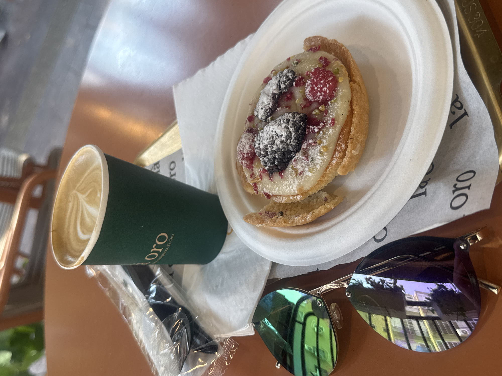 A close-up shot features a green coffee cup with a latte and foam art, alongside a pastry on a white paper plate. The pastry is topped with white frosting, powdered sugar, raspberries, and blackberries. To the left of the plate, a pair of reflective sunglasses is visible. A plastic-wrapped utensil set is also present. The items are placed on a brown table. In the background, a blurred chair and a hint of the outdoors suggest an outdoor seating area. The overall scene is set at a cafe, possibly in Ramat Gan, Israel, as suggested by the presence of what appears to be Israeli scenery reflected in the sunglasses.