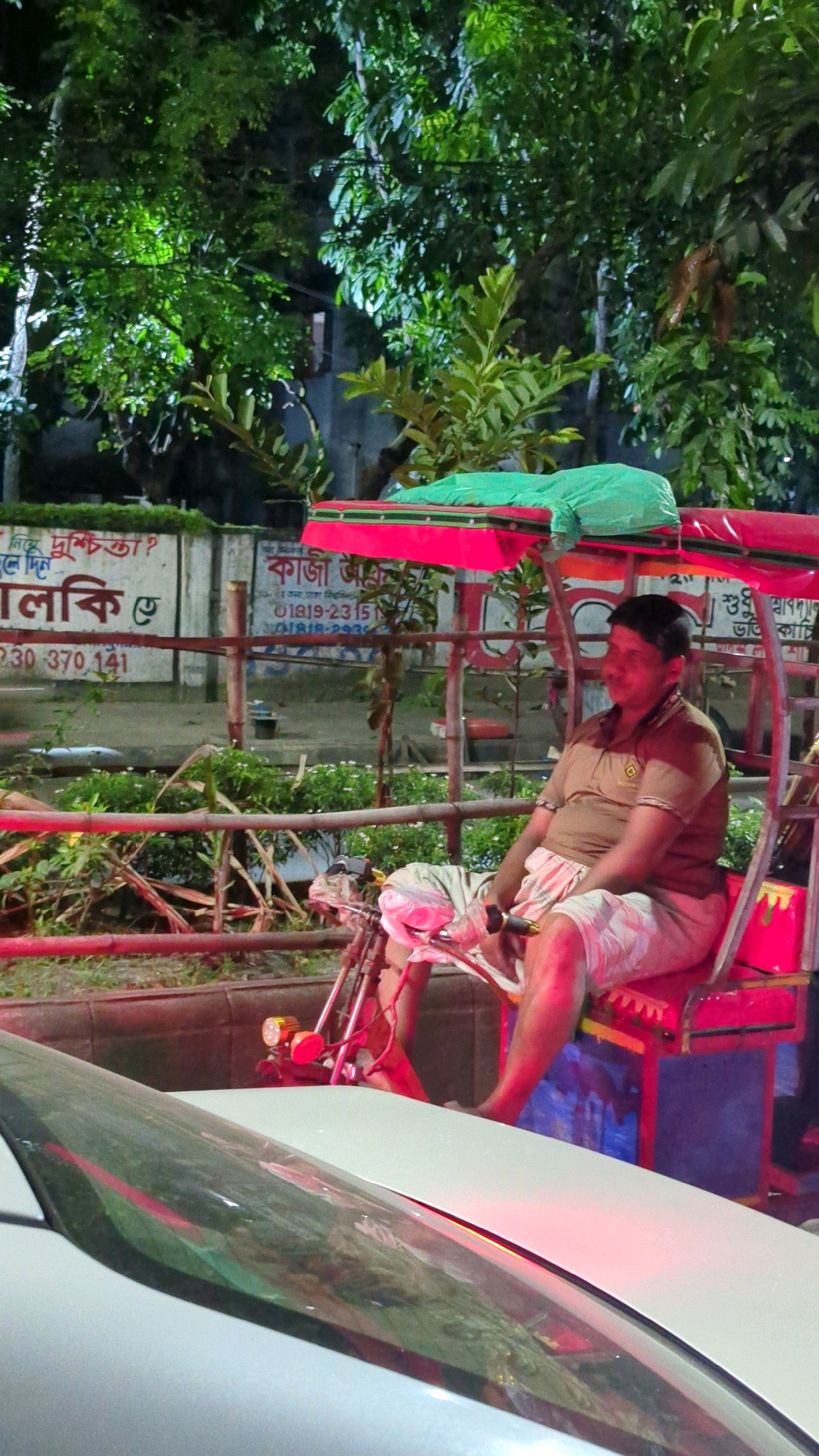 A man sits in a cycle rickshaw, the vehicle's canopy is adorned with a green cloth. The man is wearing a brown polo shirt and checkered shorts. He is positioned on the right side of the rickshaw. In the background, trees with green foliage are visible, illuminated by artificial light. A white wall with writing in Bengali is also present. The scene appears to be in Dhaka, Bangladesh, likely near a road given the presence of a white car in the foreground. The lighting suggests it is nighttime.