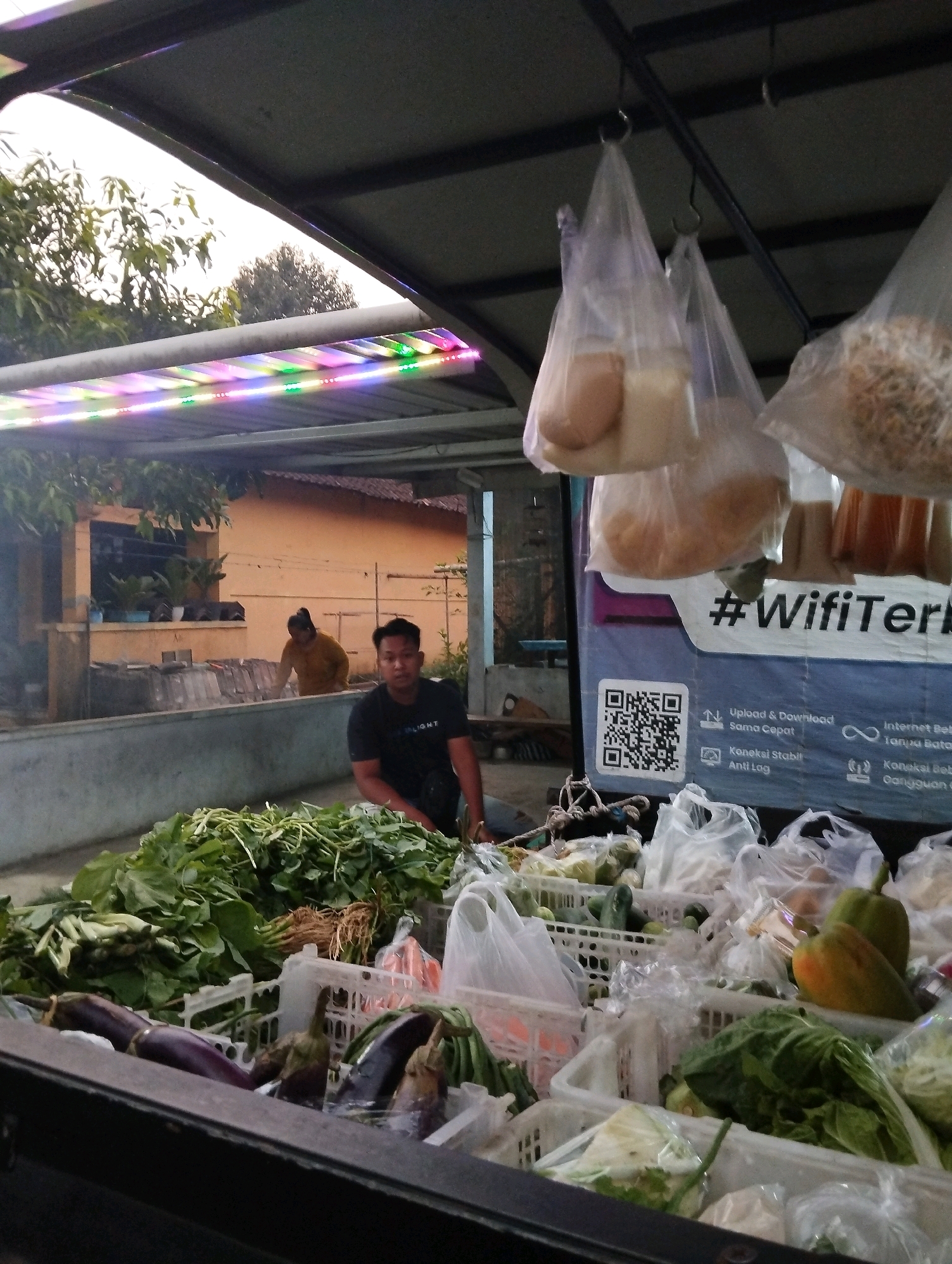This image depicts an outdoor market stall in Salatiga, Indonesia, likely near a residential area given the building in the background. The foreground is dominated by a variety of fresh produce, including leafy green vegetables, eggplants, and peppers, neatly arranged in white plastic crates. Some produce is also bagged in plastic. A young man, wearing a black t-shirt, is seated in the stall, looking towards the camera. In the mid-ground, a woman in a yellow top is tending to something behind a concrete structure. Above the stall, plastic bags containing food items are suspended. A colorful LED light strip is visible along the edge of the overhead shelter, casting a vibrant glow. A banner with text, including "#WifiTer" and a QR code, is prominently displayed behind the produce. The overall scene suggests a busy day at a local market, with goods being prepared for sale and customers potentially being served.