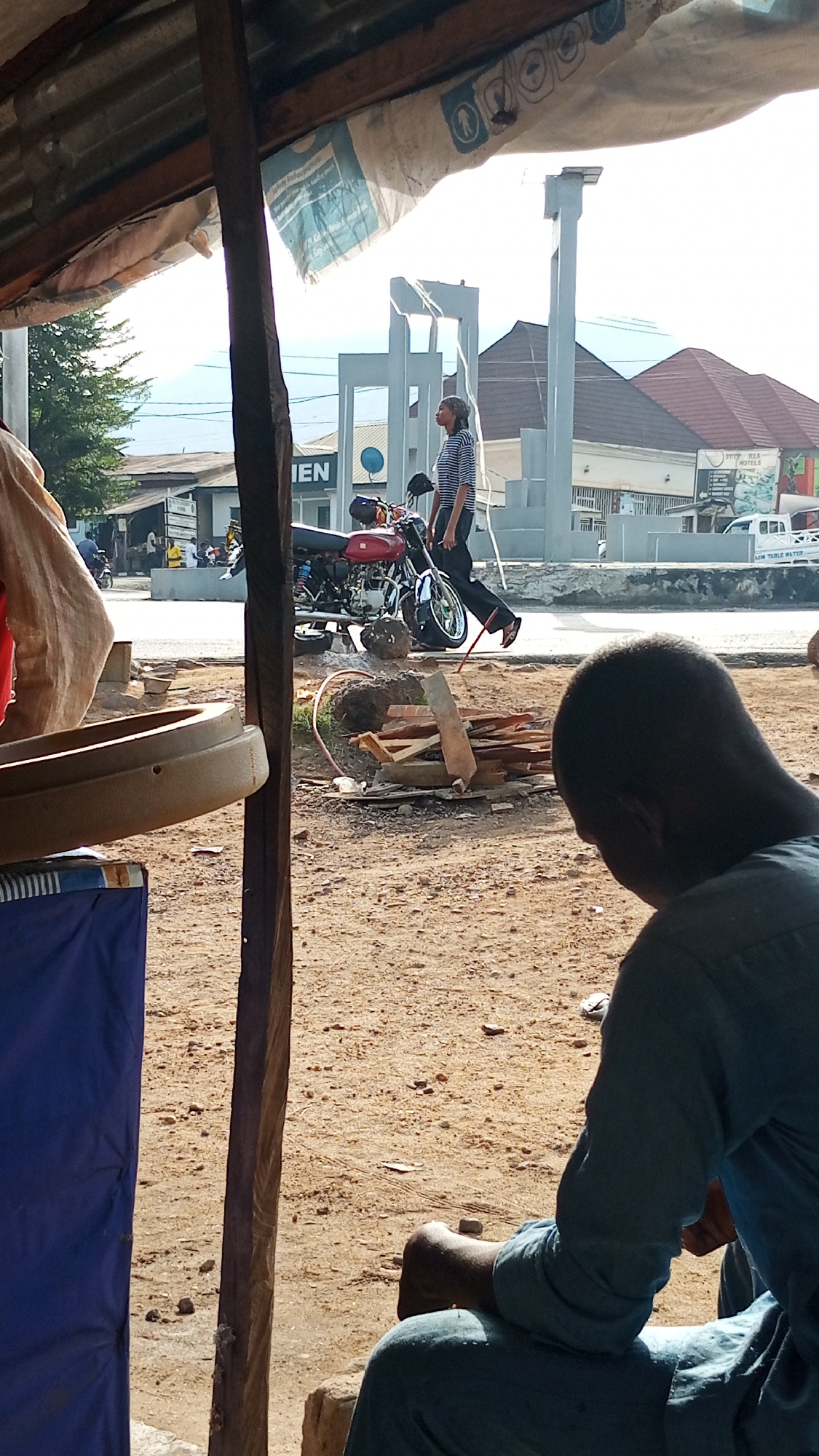 The image shows an outdoor scene in Adankolo, Nigeria, partially under a makeshift shelter. A man in a blue outfit is seated in the foreground, his back facing the camera. Through the opening of the shelter, a red motorcycle is parked next to a pile of firewood and debris. A woman wearing a striped shirt and dark pants is walking near the motorcycle. In the background, there's a building labeled "IEN" and a white vehicle. Further in the background, there's a concrete structure and a building with a reddish roof. The ground is sandy.