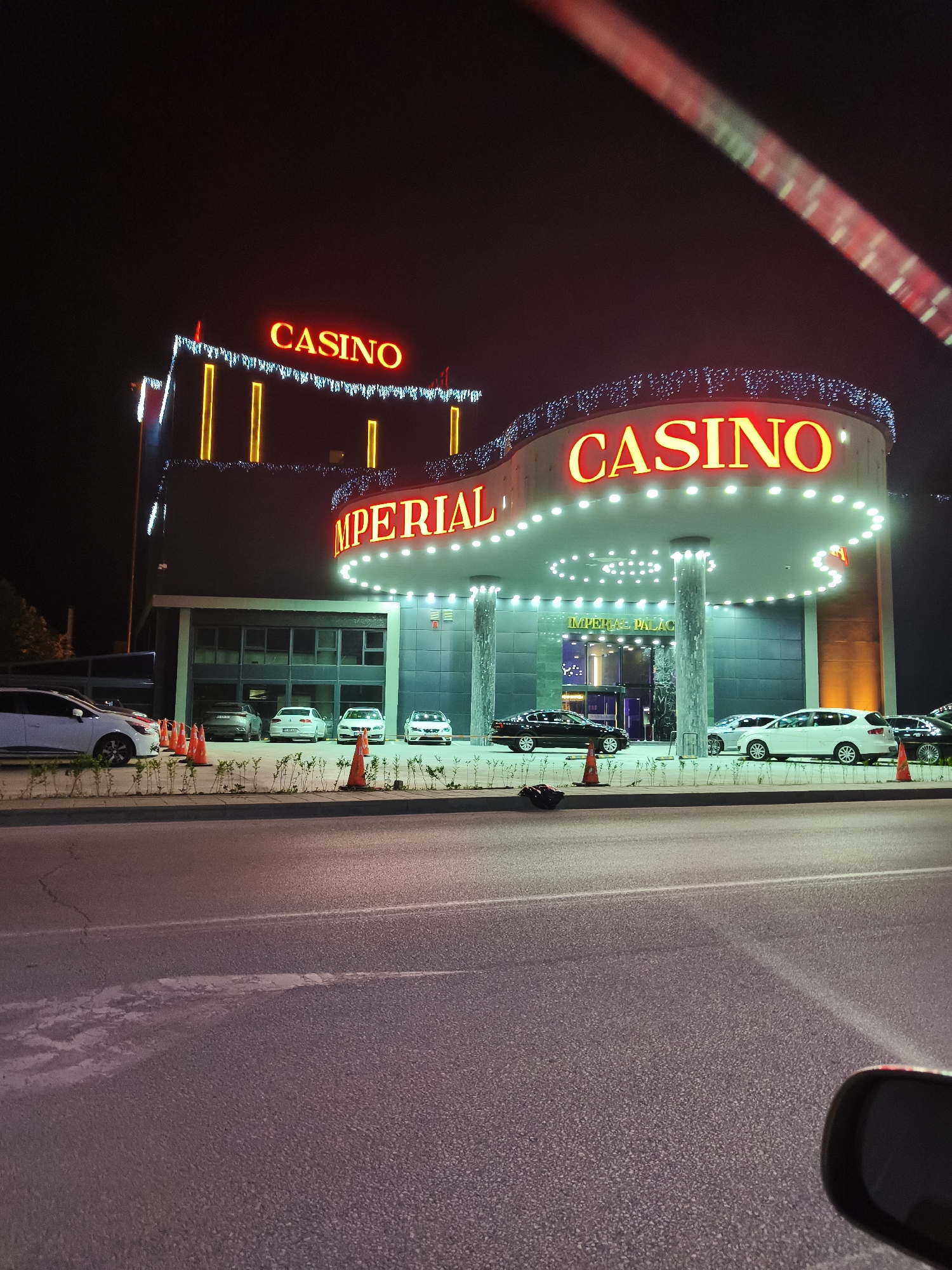 The image captures a vibrant night scene of the Imperial Palace Casino in Svilengrad, Bulgaria, viewed from the interior of a vehicle. The sky is dark, highlighting the brightly illuminated casino building.
The casino features two prominent, large red neon signs spelling "CASINO." The main entrance area is adorned with numerous small white lights outlining its curved canopy, which also has recessed downlights. Below the main "CASINO" sign, the word "IMPERIAL" is partially visible, and the full name "IMPERIAL PALACE" is inscribed on the glass doors of the entrance. The building's exterior is modern, with dark grey paneling and large windows on the lower level. Decorative golden-yellow vertical lights and string lights also adorn the building's facade.
Several cars are visible around the casino. Multiple vehicles, including white and dark-colored sedans, are parked in front of the building, with one black car appearing to be stopped or pulling up near the entrance. White traffic cones are placed along the curb, delineating the parking or access areas.
The photograph is taken from a low angle, showing a section of an asphalt road in the foreground with visible road markings and some surface imperfections. A small portion of a car's interior, possibly a dashboard or rearview mirror, is visible in the bottom right corner, indicating the vantage point. A faint reddish streak cuts across the upper right portion of the sky, likely a reflection or light artifact from inside the vehicle. The scene conveys an active, upscale nightspot, bustling with arriving patrons.