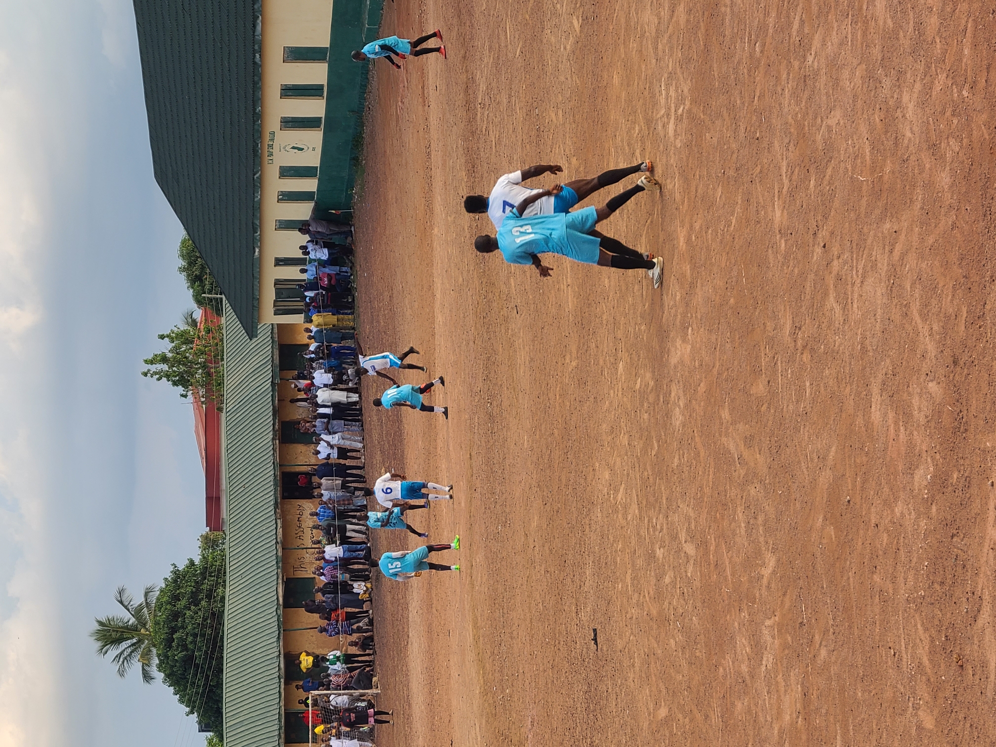 An amateur outdoor soccer match is underway on a reddish-brown dirt field under an overcast sky. The location is identified by a sign on one of the background buildings as "R.C.M. PRIMARY SCHOOL, ZANGONKATAF" in an unknown city, unknown country.
Approximately ten players are visible on the field, divided into two teams: one in light blue jerseys and shorts, and the other in white jerseys with blue numbers and trim, and blue shorts.
On the left side of the frame, near a white goal frame with netting, a player wearing a light blue jersey with the number 15 is standing, facing right. Another light blue player (number 13, back to viewer) is positioned in the lower-center, facing right, seemingly engaged with a white-jerseyed player (number 7, back to viewer) who is facing left. In the mid-ground, a white-jerseyed player with the number 6 is standing, facing left. Other players in both light blue and white jerseys are visible across the field, some in motion. A goalkeeper in a black jersey and pink shorts is visible near the left goal.
The field itself is dry, uneven, and devoid of grass or visible markings.
In the background, two single-story buildings with light yellow/tan walls and dark green corrugated metal roofs line the far side of the field. The building on the left has "THIS IS ASSEMBLY AREA" written in black on its wall and is partially obscured by a tall palm tree. The building on the right clearly displays the "R.C.M. PRIMARY SCHOOL, ZANGONKATAF" signage and a crest. Approximately 70-100 spectators, primarily adults, are gathered along the perimeter of the field in front of these buildings, observing the game.
