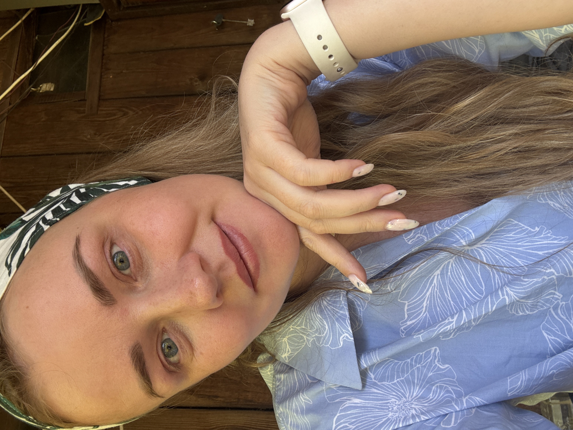 A woman is photographed in a head-and-shoulders shot. She wears a blue floral-patterned blouse and a patterned headscarf. Her hair is long and light brown. She is wearing a smartwatch with a light-colored band. Her nails are manicured. The background consists of a wooden surface and, in the upper left corner, visible wires and a window. There is no indication that this image was taken in Chaikovsky, Russia.