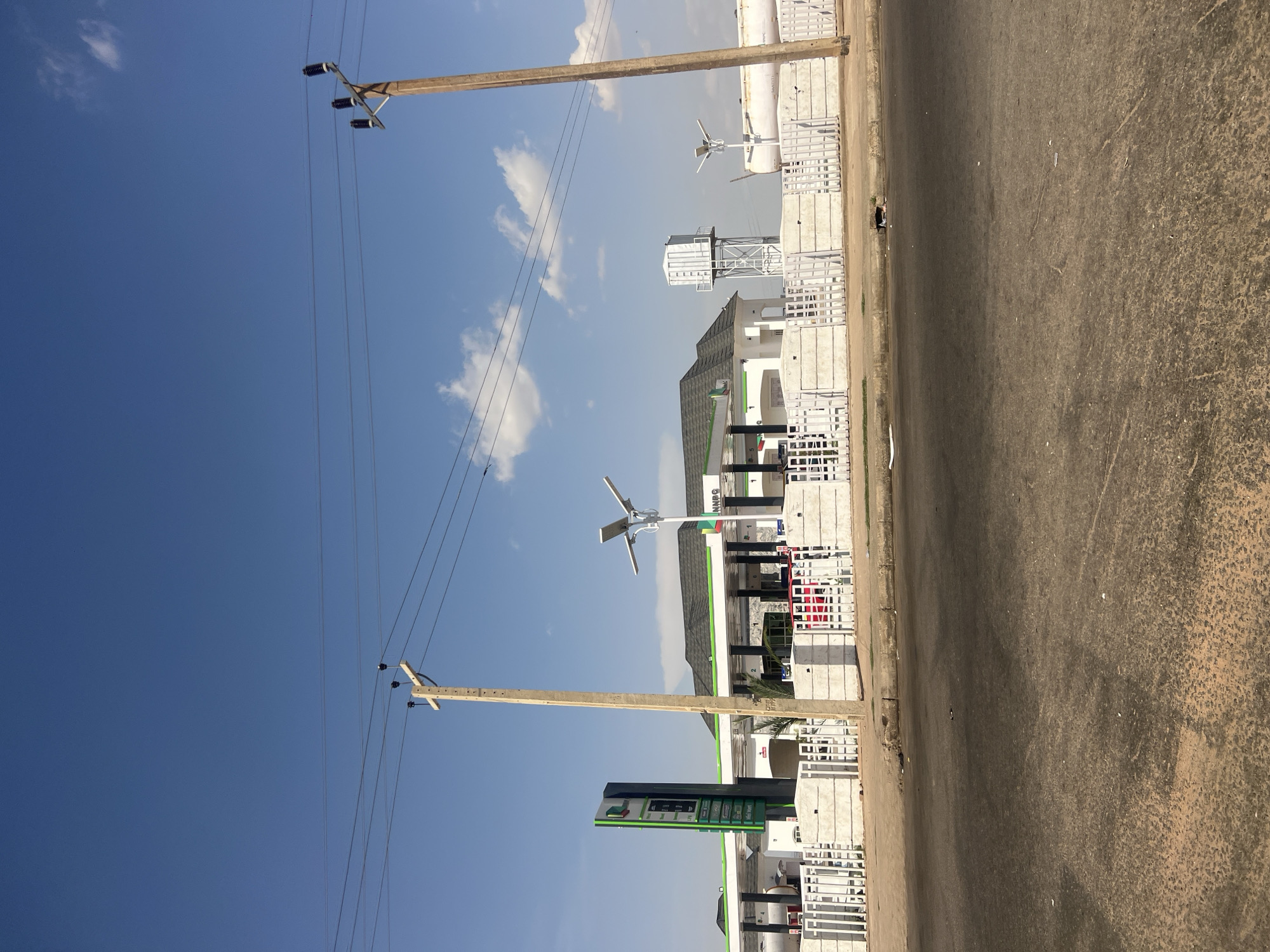 The image depicts a daytime street view of a petrol station in Gombe, Nigeria, under a clear blue sky with scattered white clouds. In the foreground, a dark asphalt road with minor debris is visible, bordered by a concrete curb and sidewalk. Two tall, light-colored wooden utility poles with multiple black power lines stretch across the upper portion of the frame, one on the left and one on the right.
Behind the sidewalk, a white concrete and picket fence encloses the petrol station premises. The station features several white buildings with dark gray roofs and green trim, appearing to be dispensing areas under canopies. A tall, green and white vertical sign, likely displaying fuel prices, is prominent on the left side of the station. The letters "NNPC" are visible on the front fascia of one of the canopies. Modern streetlights with solar panels are integrated into the station's infrastructure, with one distinctive three-panel light stand positioned centrally. To the right, a tall, white square water tank on a metal support structure stands behind the station buildings, and further right, a large white horizontal storage tank is visible. No people or vehicles are clearly identifiable within the scene.