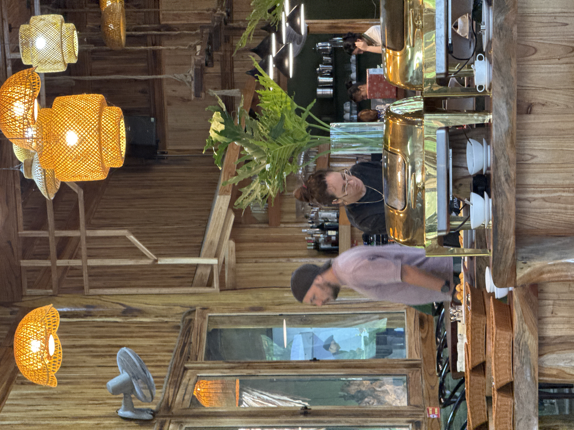 The image shows the interior of a restaurant in Pù Luông Commune, Vietnam. The restaurant has a rustic design with extensive use of natural wood. Hanging from the ceiling are multiple woven lampshades in warm, orange tones, casting a soft glow. A wall-mounted fan is visible on the left. In the foreground, a buffet setup features two large, polished brass chafing dishes, suggesting a meal service is underway or has recently concluded. Several woven baskets filled with food items are arranged on a wooden counter.
In the midground, a man wearing a purple t-shirt and a dark beanie is standing to the left, looking towards the right. A woman with her hair in a bun, wearing glasses and a dark top, is seated behind the buffet counter, appearing to be engaged with something on the counter. A large potted plant with broad green leaves is positioned in a clear glass vase on the counter between the woman and the chafing dishes, adding a touch of natural greenery to the scene. Behind the counter, shelves are stocked with bottles, glassware, and other items. Further in the background, more people are visible, some seated and others standing, suggesting other patrons or staff are present. The overall impression is of a relaxed and natural dining environment.