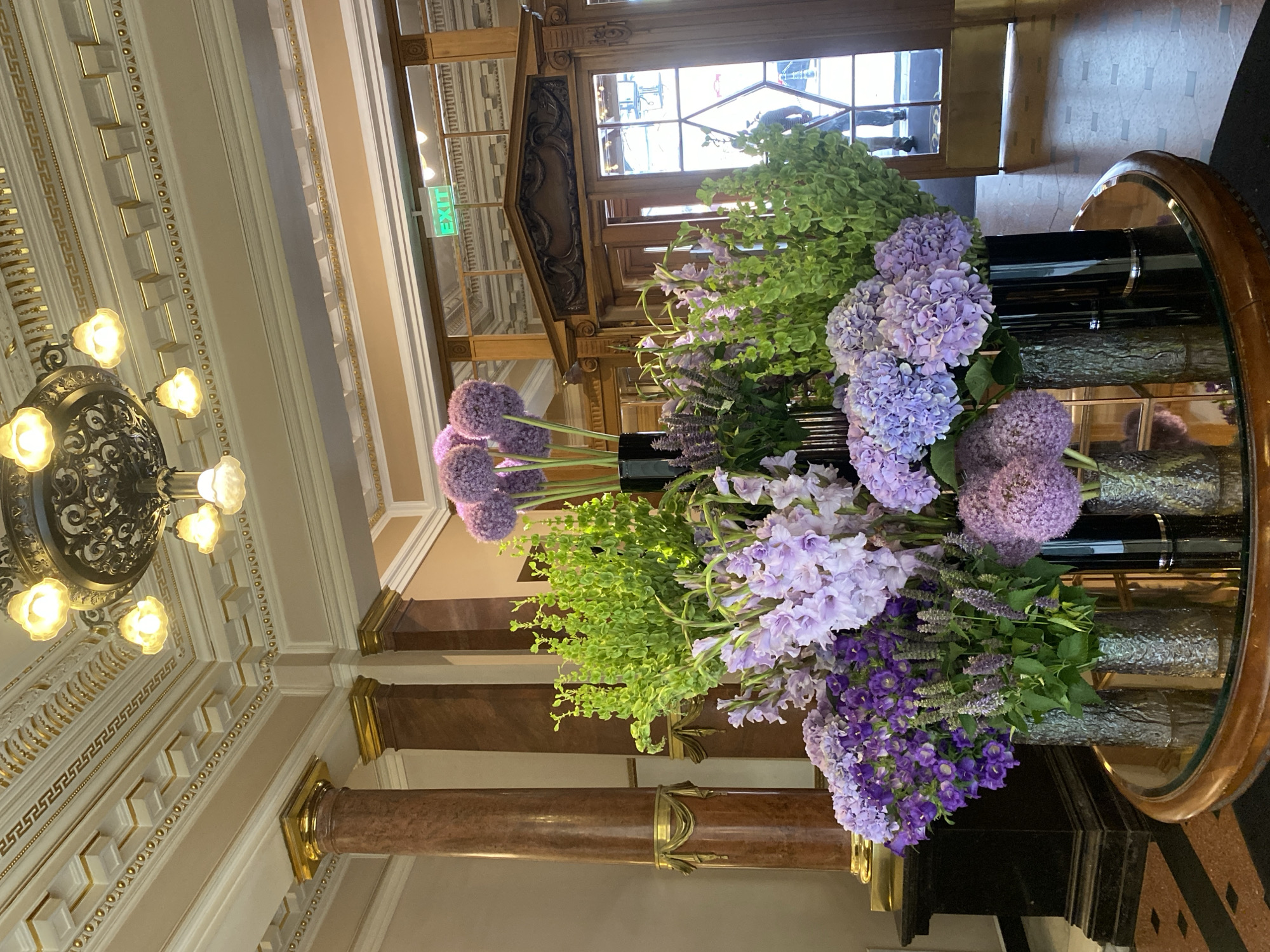 The image depicts a grand hotel lobby interior, likely in Saint Petersburg, Russia, characterized by opulent classical architecture. Dominating the foreground is an elaborate floral arrangement featuring a variety of purple and green flowers, including hydrangeas, alliums, and what appear to be gladioli. These flowers are displayed in several dark, cylindrical vases of varying textures. The arrangement sits on a circular, reflective surface, possibly a glass-topped table with a wooden rim.
In the background, the lobby showcases marble-clad columns with gold accents, a highly decorative ceiling with intricate moldings and a large, ornate chandelier with multiple lights. To the right, a set of double wooden doors with glass panels provides a view into an adjacent area. An illuminated green "EXIT" sign is visible above the doors. The floor is a mosaic of dark and lighter tiles, with a section of patterned carpeting visible on the left. The overall impression is one of luxury and classic elegance.