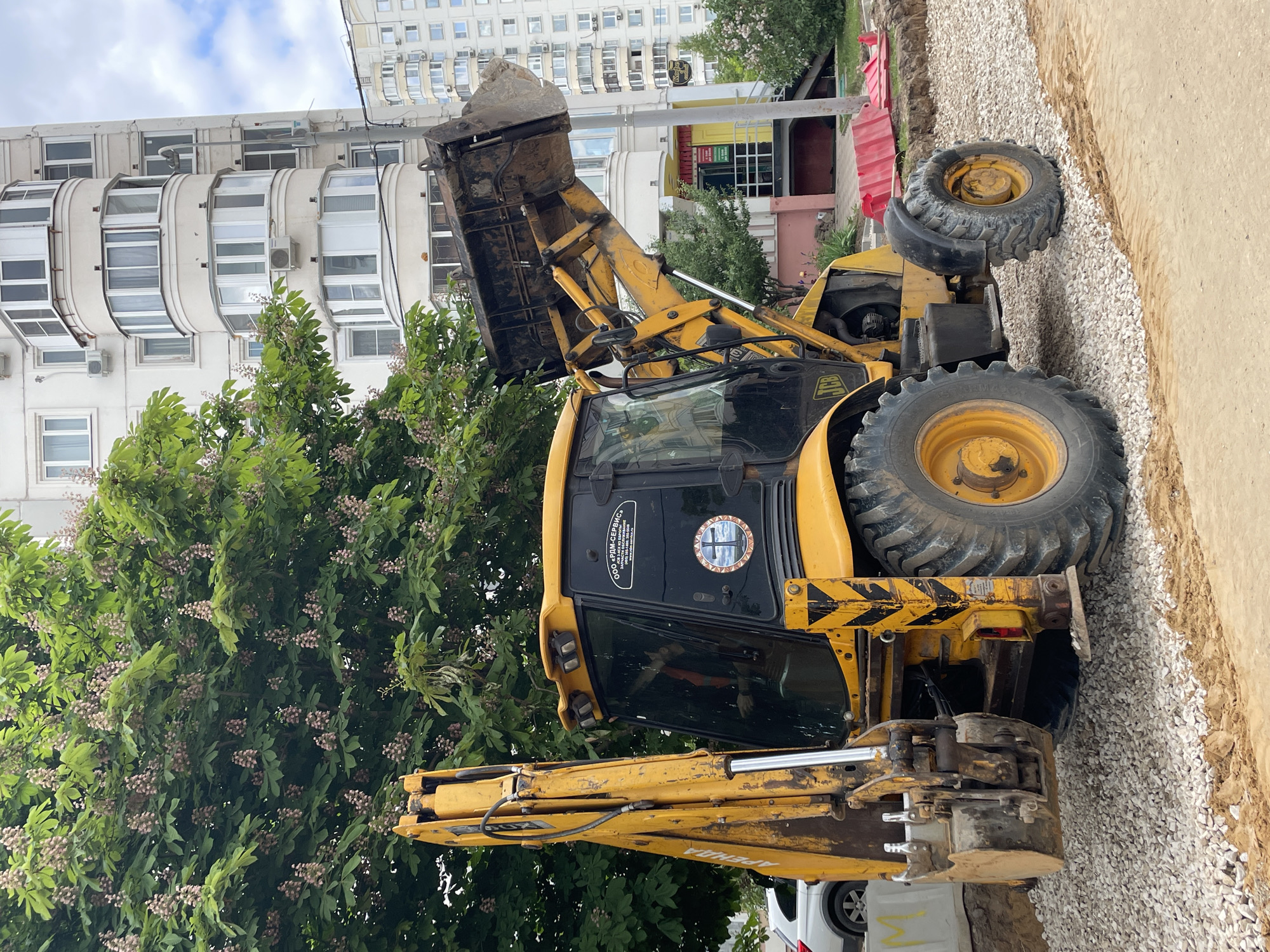 A yellow JCB backhoe loader is parked on a construction site in Moscow, Russia. The backhoe arm is extended and positioned behind the vehicle. The bucket is raised and angled upward. Construction materials, including gravel and dirt, surround the backhoe. A row of red and white safety barriers are visible. In the background, there are buildings, including residential apartments. The sky is overcast with scattered clouds. Trees with green foliage are visible near the backhoe. The backhoe cab has a sticker on its back window with text and logos in Russian. A partial view of a white vehicle is on the left.