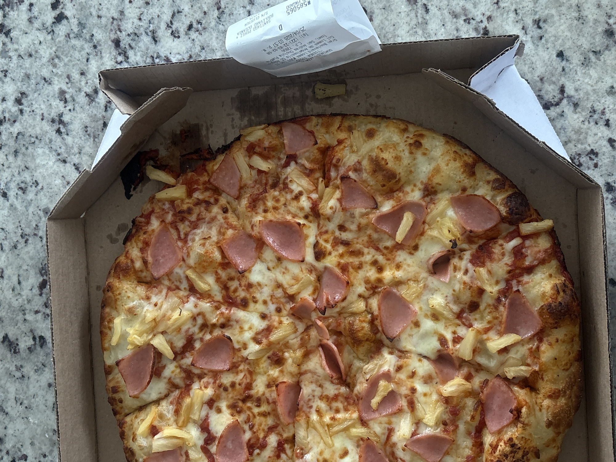 The image shows a partially eaten Hawaiian pizza in a cardboard box. The pizza has a crust, cheese, tomato sauce, ham, and pineapple toppings. A receipt from Domino's Pizza is on top of the box. The receipt indicates that the order was placed at store number 11575, located on Gran Boulevard Lomas #301 in Lomas de Angelopolis CC Town, San Andres Cholula Puebla, C.P. 72830, Mexico. The transaction date is 25/04/2026, 03:57 p.m. The receipt shows the store number is 585065. The background is a countertop with a speckled design.