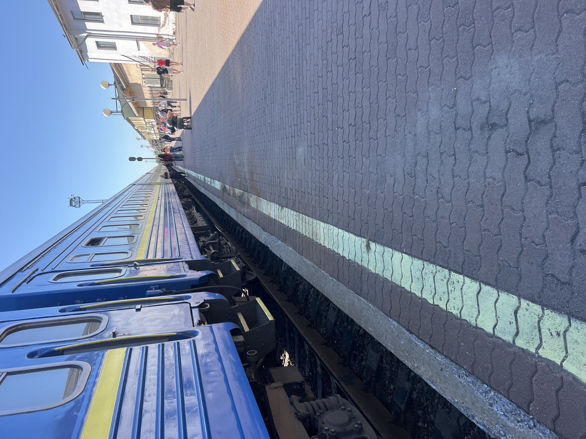 An exterior shot captures a blue and yellow train stopped at a station platform under a clear blue sky. The train occupies the left side of the frame, its blue carriages with yellow stripes running lengthwise. The platform, paved with interlocking brick pavers in shades of dark gray and brown, stretches to the right, marked by a bright yellow safety line. Several people are visible on the platform, some walking, others standing, and one individual is pushing a luggage cart. In the background, a white building with multiple windows and a longer, cream-colored building with yellow accents are situated along the platform. A signal light is mounted on a pole near the train, and a cellular tower is visible in the distance on the left. The scene is consistent with a train station in Ivano-Frankivsk, Ukraine.