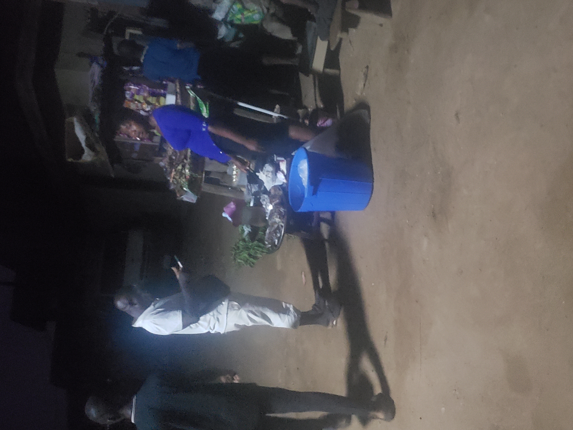 Here is a detailed description of the image:
**Location:** Alalubosa, Nigeria.
**Scene:** The image appears to be taken outdoors in what looks like a market or roadside stall environment at night.
**People:** Several people are visible, though visibility is limited.
* One person is wearing a blue shirt and appears to be interacting with the stall.
* Another person, closer to the camera, is wearing light-colored pants and a white shirt. They appear to be holding a phone or similar device.
* A third person, mostly in shadow, stands near the edge of the frame, wearing a dark jacket.
**Objects:** Key objects in the scene include:
* A roadside stall or display, stocked with various items.
* A blue plastic bucket sitting near the stall.
* Vegetables or produce on display.
**Actions/Interactions:**
* A person in the blue shirt appears to be shopping or inspecting items at the stall.
* The person in the white shirt is holding their phone and seemingly observing the scene.
**Environment:** The environment looks like a typical informal market area, likely at night. The ground is dirt or pavement. Lighting is poor.