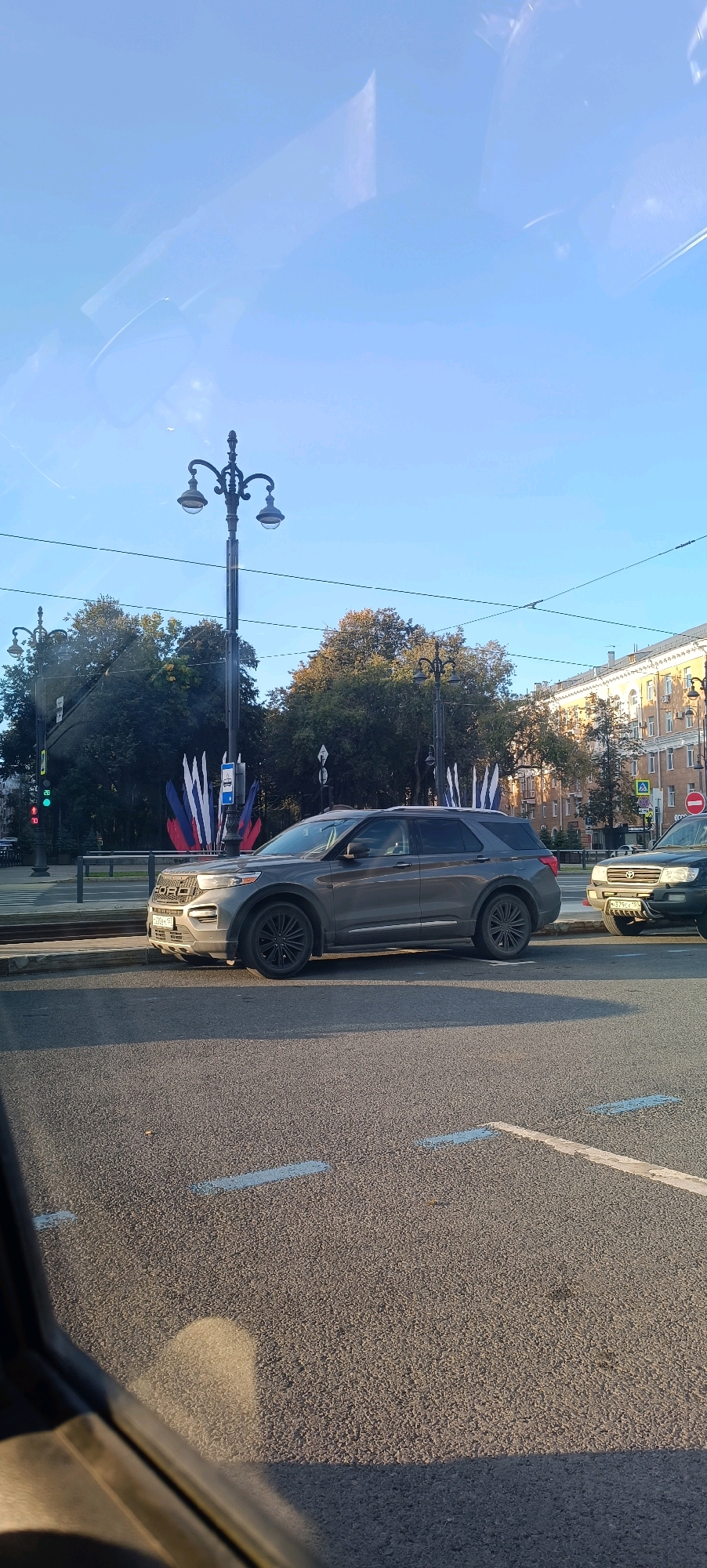 In Perm, Russia, on a clear day, a gray Ford Explorer is parked on the side of a city street, facing right. The vehicle is positioned on the asphalt road, near a curb. Behind the Ford, a black SUV is also parked. In the background, trees with autumnal foliage stand, and a multi-story building with a light-colored facade is visible. A street lamp with two lights is prominent in the middle ground. Several flags, seemingly in red, white, and blue, are displayed behind the Ford Explorer. Traffic lights are visible on the left side of the image. The scene appears to be a street in a city, with buildings, trees, and vehicles present.