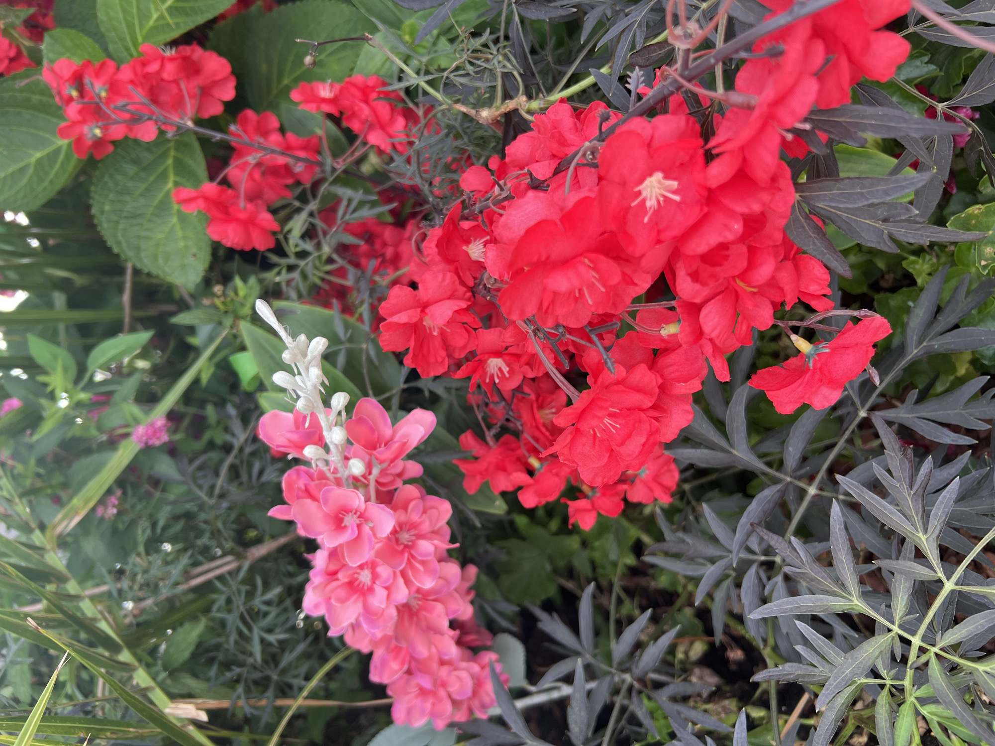 A vertical close-up shot shows clusters of bright red and pink artificial flowers and dark green foliage. The flowers appear to be made of fabric and are arranged on thin stems. The pink flowers are located in the upper left of the frame, while the red flowers are in the center and right. Dark, leafy plants with pointed leaves are visible in the lower portion of the image. In the background, there are blurred green leaves and hints of other pink flowers. The lighting is bright and even, suggesting an outdoor setting during the day. There are no people or animals present.