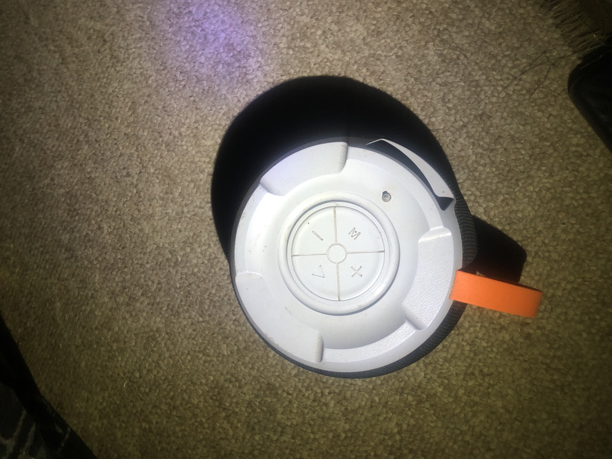 A portable Bluetooth speaker with a white top is resting on a brown carpet. The speaker has a circular control panel with buttons labeled with symbols, including a triangle, a plus sign, a minus sign, and the letter "M". An orange loop is attached to the side of the speaker. The lighting in the scene suggests it is indoors, possibly in a room in Jalingo, Nigeria. There are no people or animals visible in the image.
