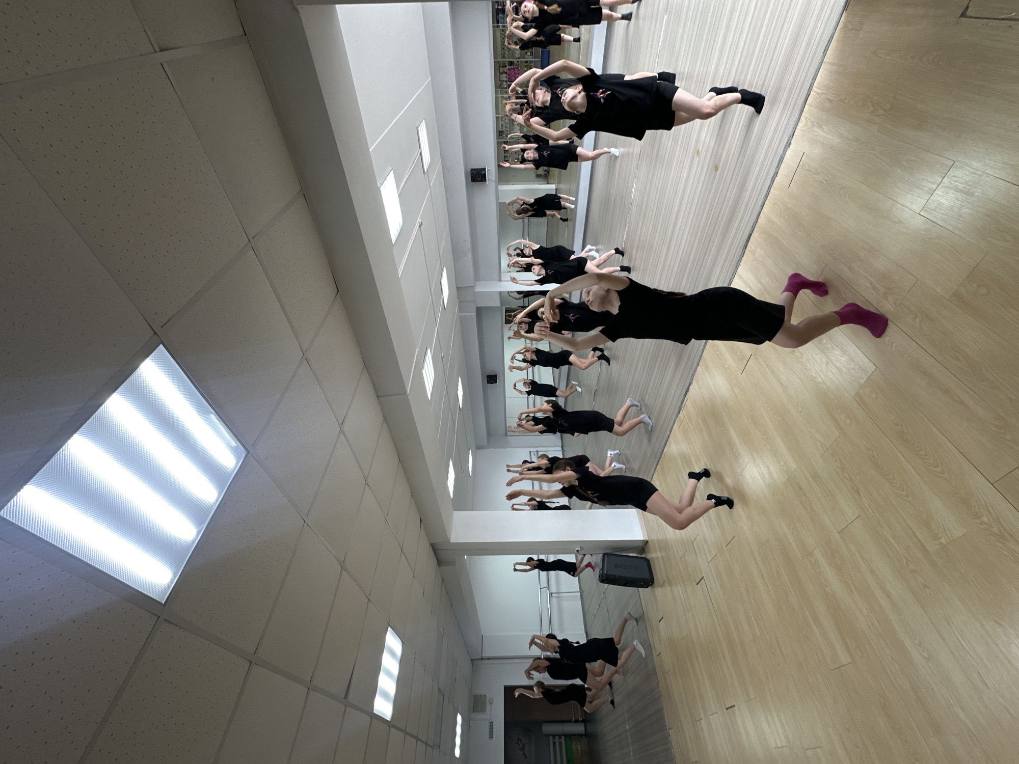 This image captures an indoor scene within a dance or gymnastics studio in Votkinsk, Russia, where a group of young individuals is practicing a synchronized routine. The studio features a light-colored wooden floor and an extensive mirrored wall that reflects the participants, creating an illusion of a larger group. The ceiling is composed of acoustic tiles, fitted with bright rectangular fluorescent light fixtures that illuminate the space.
The main subjects are approximately ten to fifteen young individuals, primarily girls or young women, uniformly dressed in black t-shirts and black shorts. They are arranged in a line, facing the mirrored wall, executing an identical pose: bodies gently arched, arms extended forward and slightly upward, and legs bent at the knee with feet lifted off the ground, resembling a "superman" or "flying" position. Their reflections in the mirrors show them facing the viewer, displaying expressions of concentration and engagement. While most wear black socks, some can be seen with white or vibrant pink socks.
Visible objects in the studio include a horizontal ballet barre positioned behind the reflected figures. A black portable speaker, clearly branded with the "SONY" logo, rests on the floor to the right of the reflected group. On the wall opposite the mirrors, a green sign with the Russian word "ВЫХОД" (meaning "EXIT") is mounted above a doorway. The bright, artificial lighting suggests an active practice session.
