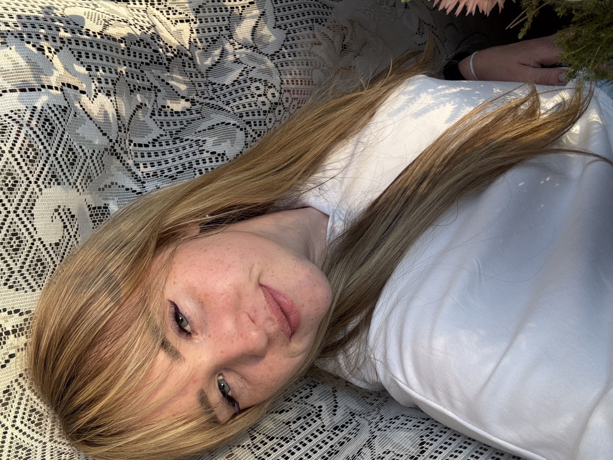 A young woman with blonde hair and freckles is lying down, looking at the camera. She is wearing a white hoodie. She has on dramatic purple eyeliner and her hair is long and blonde. She is lying on a white lace-like fabric. The lighting suggests it might be outdoors, with shadows from foliage dappling her face and clothing. The environment appears to be in the city of Марковский, Russia, based on the context provided.