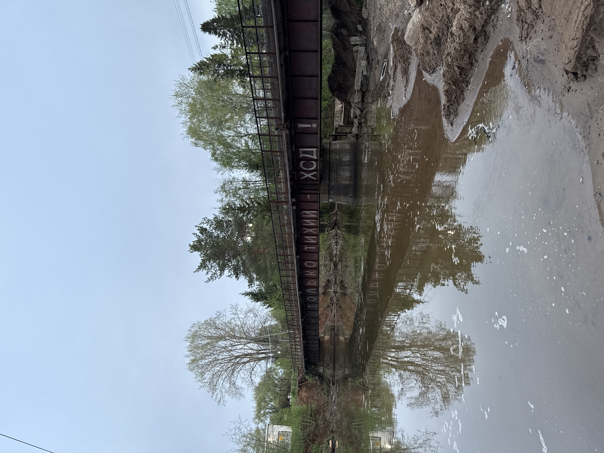 A long, horizontal photograph shows a narrow river and a bridge spanning it. On the bridge's side, "ТОЛЬКО ТИХИЙ ХОД!" is painted in white Cyrillic letters. The water reflects the bridge, trees, and sky, which is overcast. The riverbanks are earthen, and on one side, there are some rocks and sand. Trees are visible along the riverbanks and above the bridge. Power lines are visible in the sky. The city is unknown.