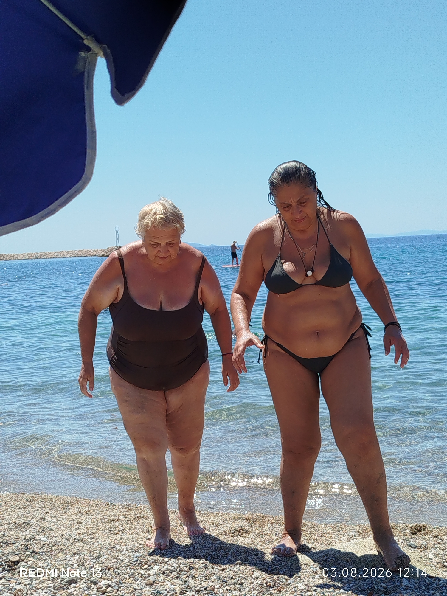 Two women are walking out of the sea onto a pebble beach. The woman on the left wears a dark brown one-piece swimsuit. The woman on the right wears a dark bikini. Both women have wet hair and appear to be exiting the water on a sunny day. The sea is calm and blue, and the sky is clear. A partial view of a blue umbrella is visible in the upper left corner of the image. In the background, a person is seen on a paddleboard in the water, and a lighthouse-like structure is visible on a distant jetty. The lower portion of the image contains a watermark with "REDMI Note 13" and a date stamp of "03.08.2026 12:14". The location is the coast of Sithonia, Greece.